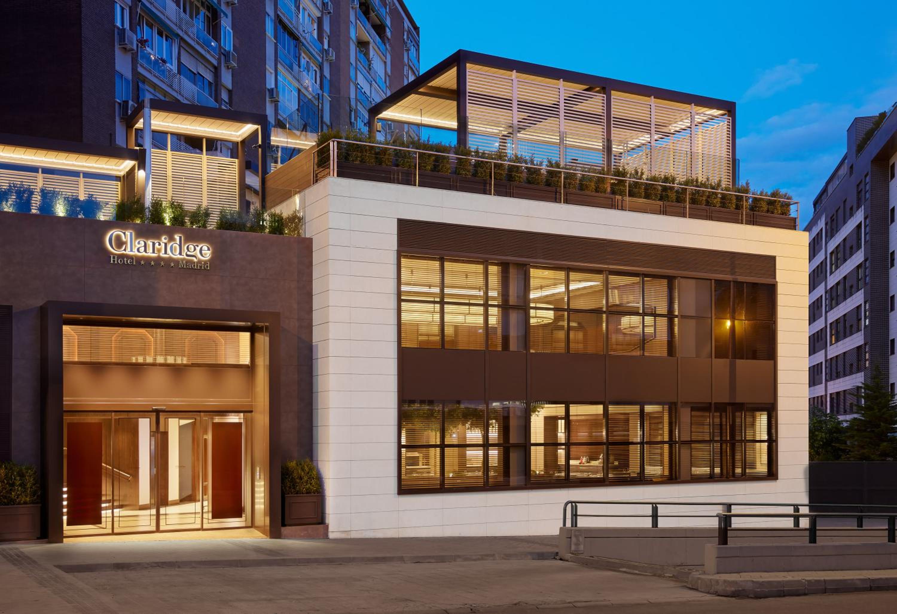 Illuminated street facade of Hotel Claridge at dusk, with large windows and a covered rooftop terrace.