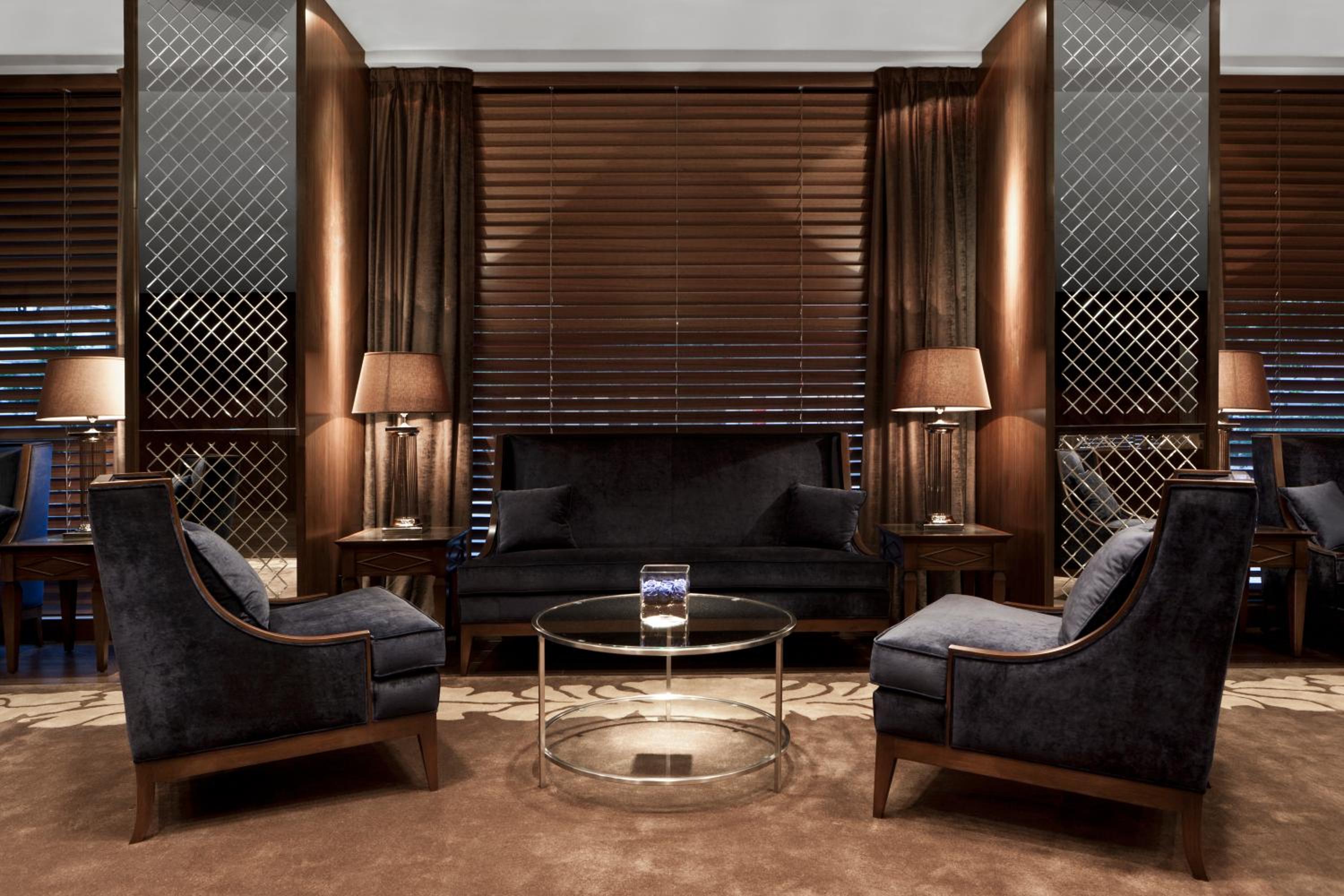 Indoor lounge with dark upholstered armchairs, glass coffee table, warm lamps, and dark wood wall panels.