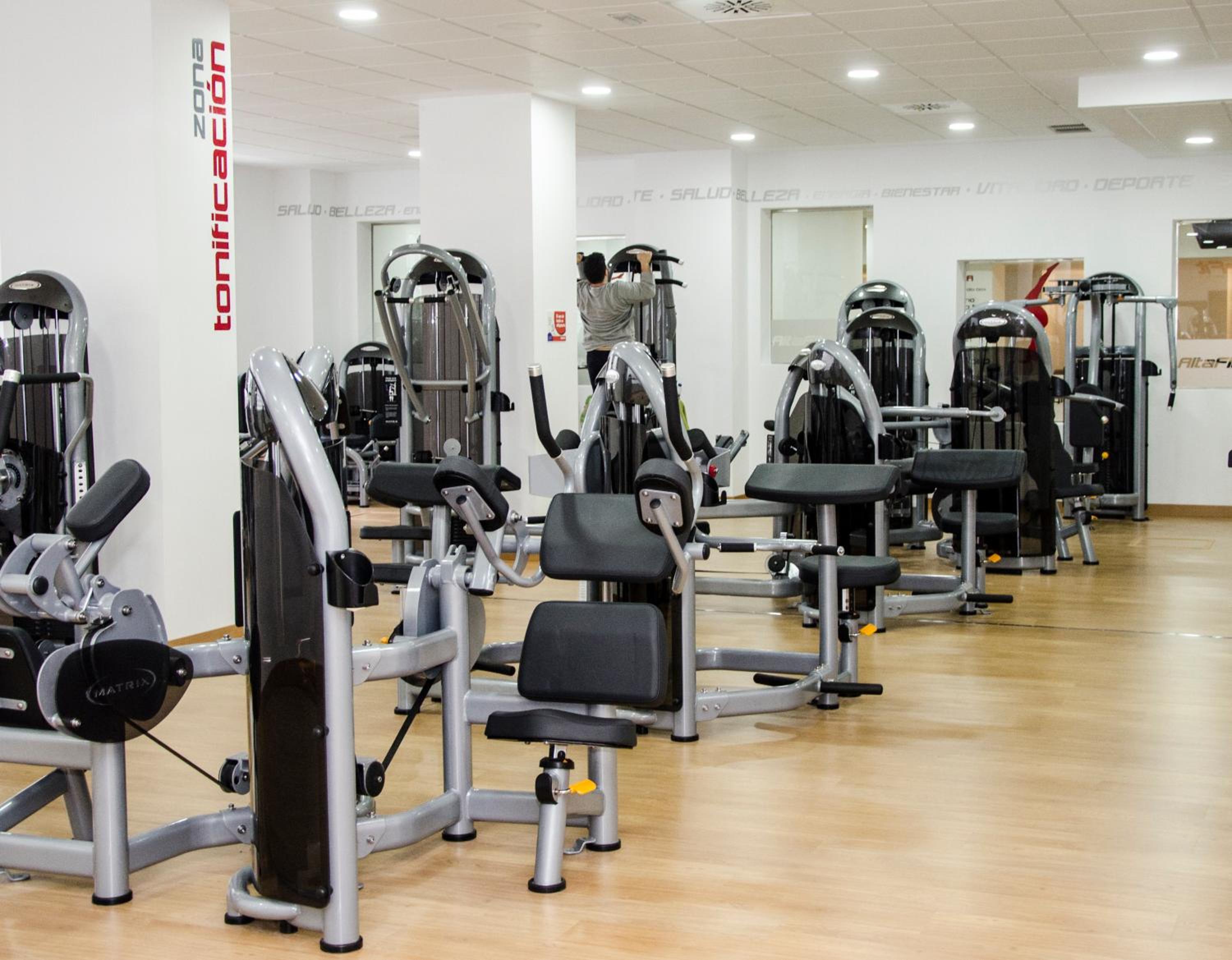 Fitness center lined with weight machines along light wood flooring under overhead lighting.