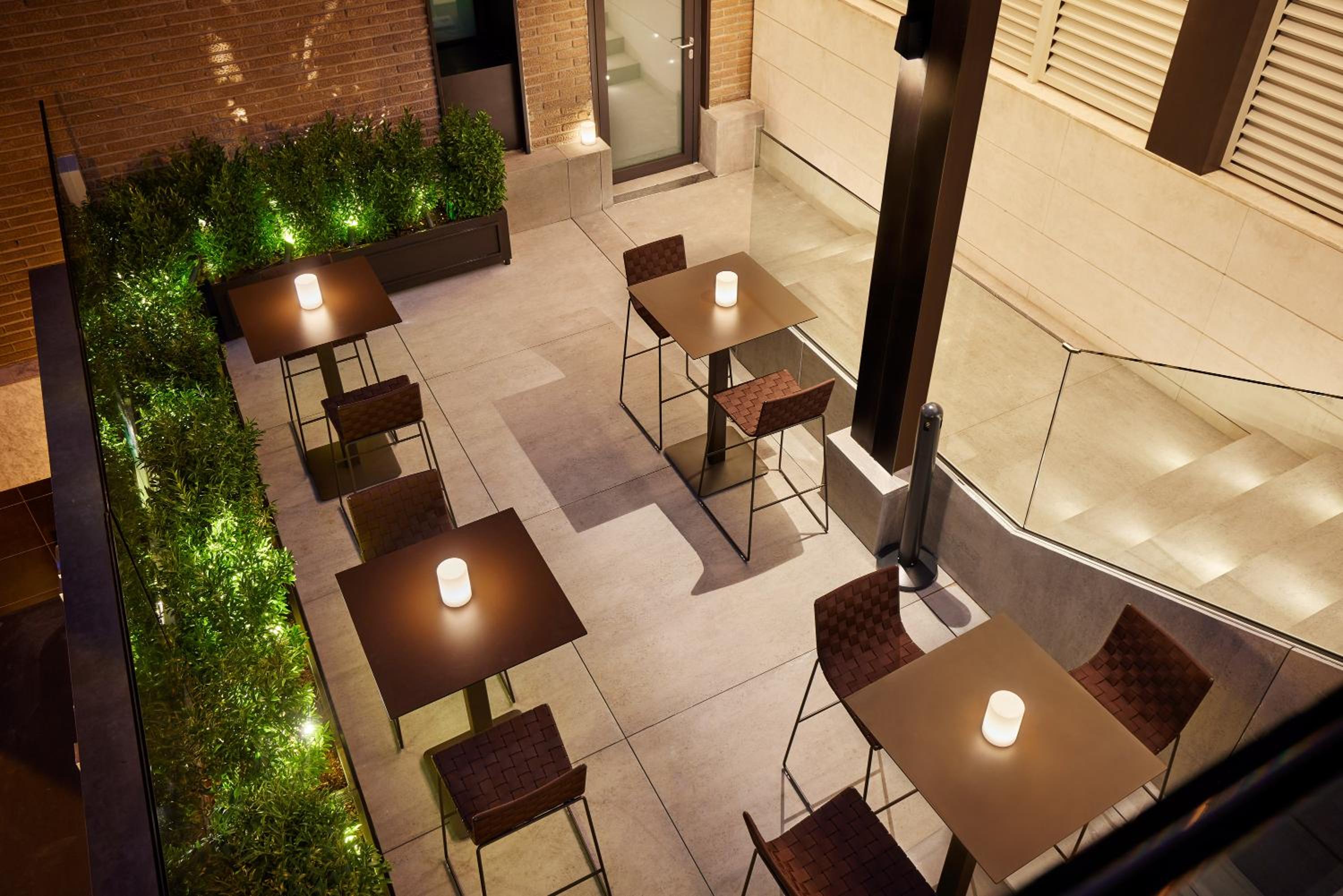 Overhead view of an outdoor patio with square cocktail tables, glowing lanterns, and boxwood planters.