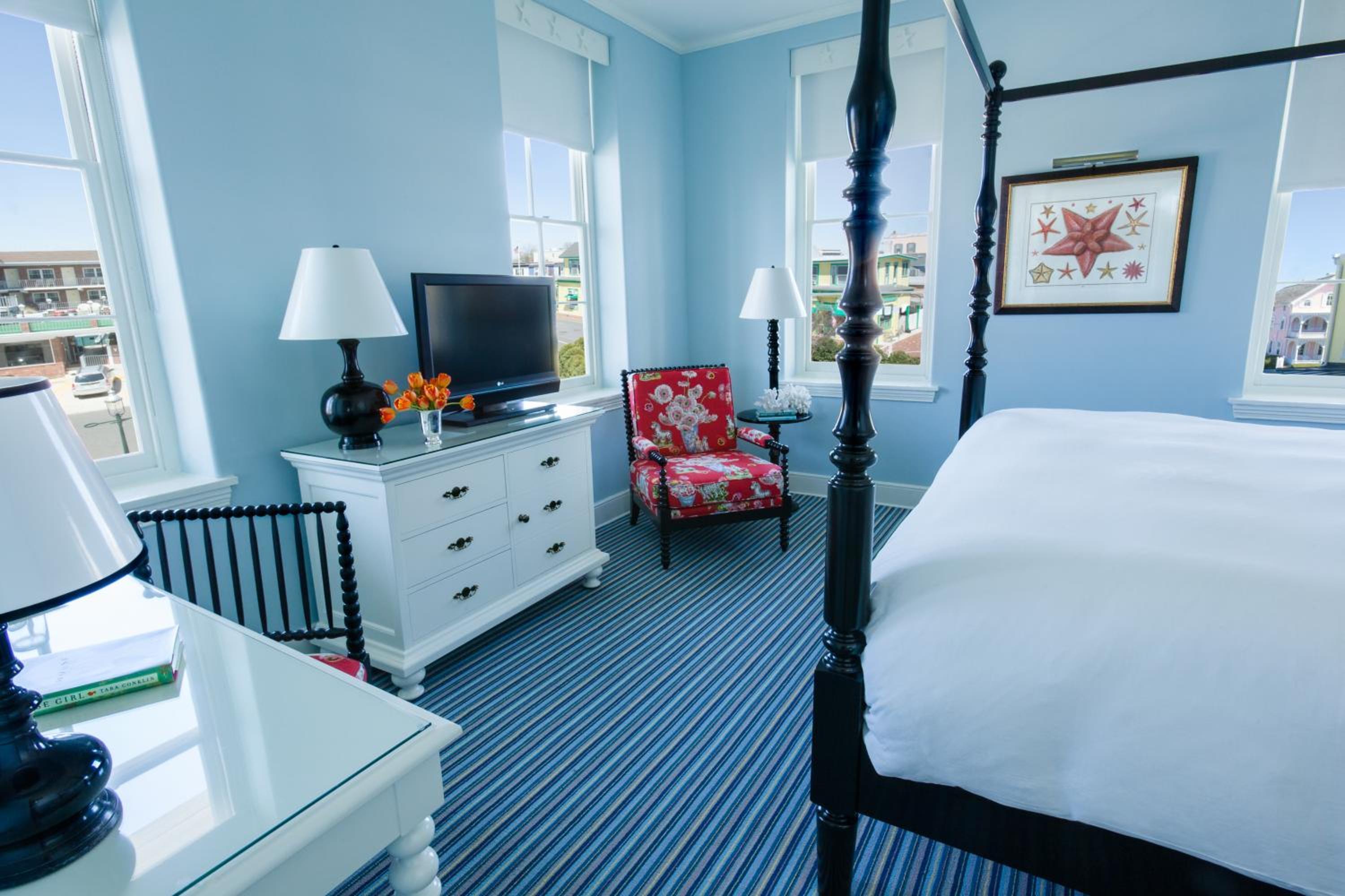 A bedroom with pale blue walls, striped carpeting, a dark canopy bed, and a red floral armchair.