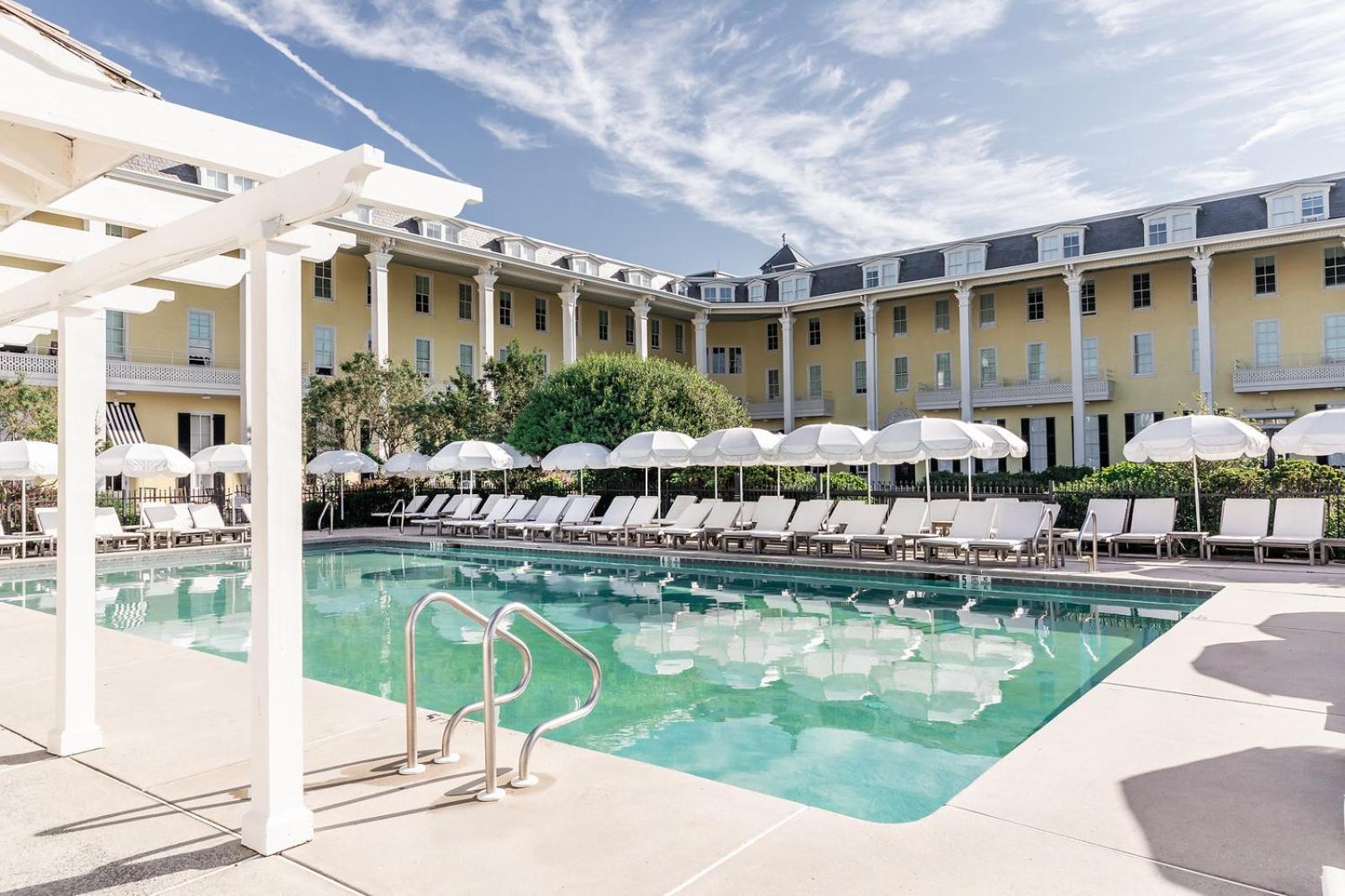 An outdoor swimming pool lined with white lounge chairs and fringed umbrellas before a yellow building.