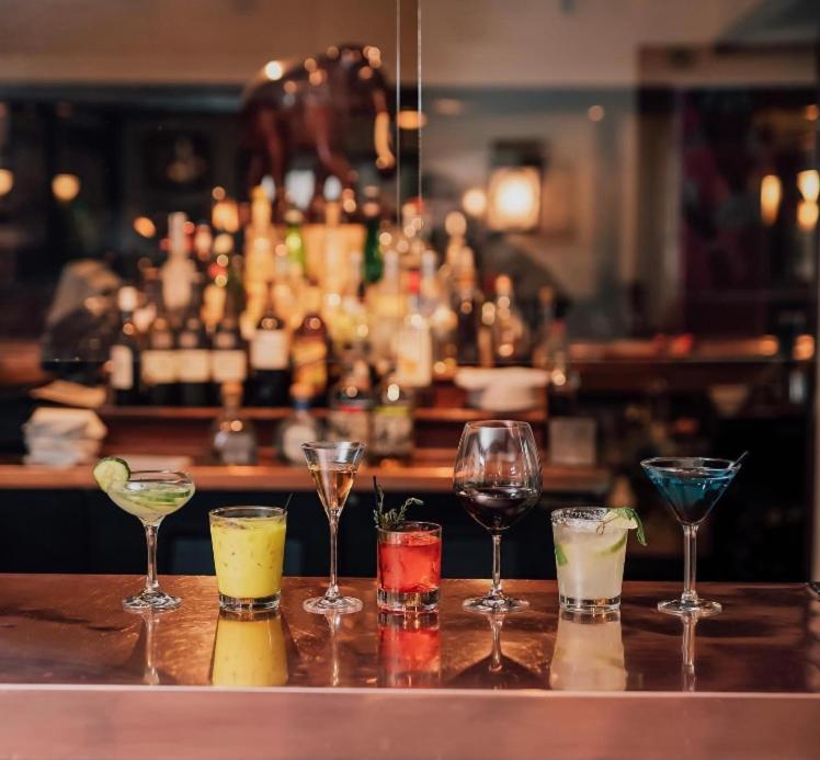 Colorful cocktails and wine glasses lined up across a polished bar top in front of illuminated liquor shelves.