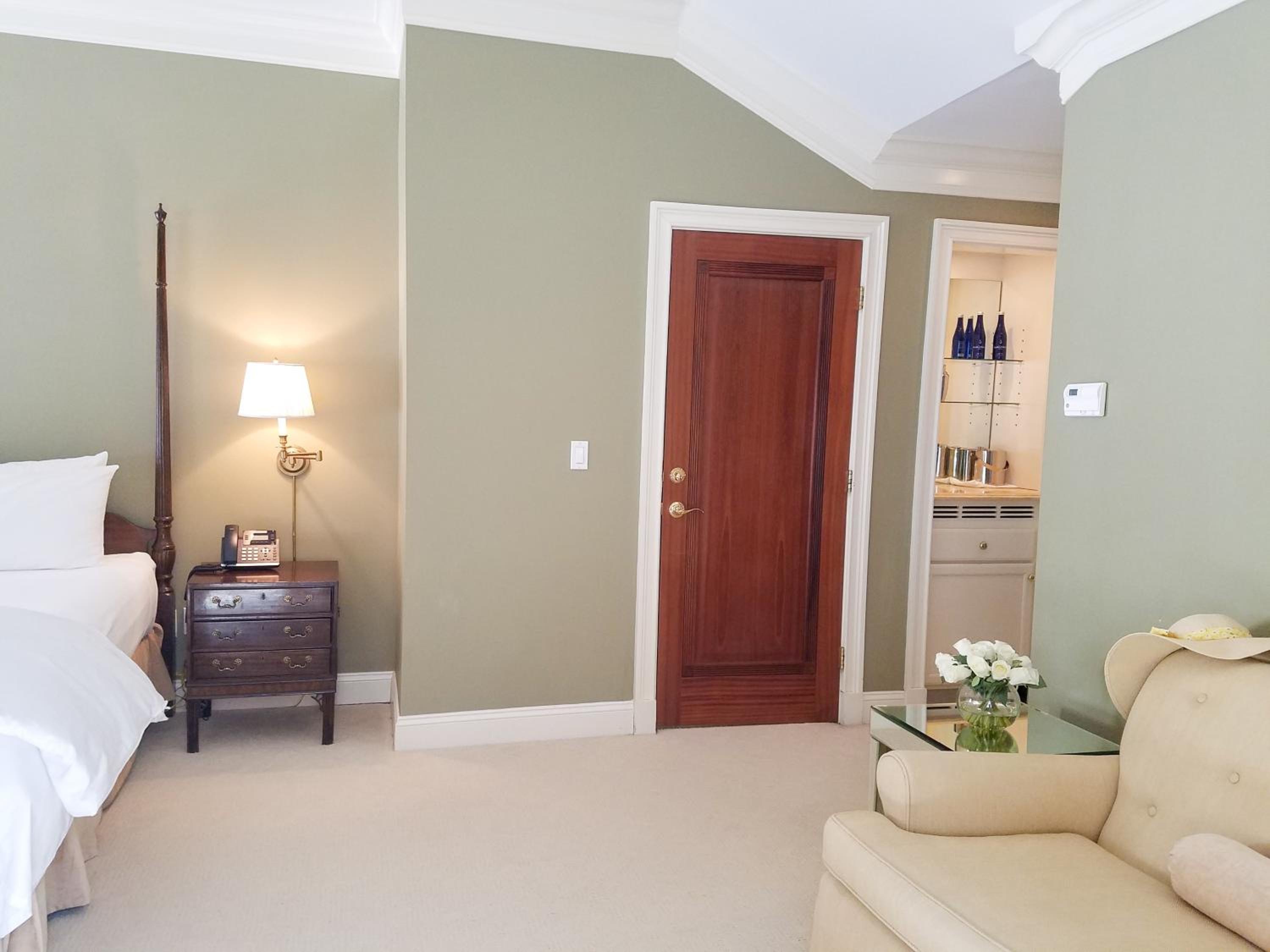 A bedroom with pale green walls, an armchair, a bedside lamp, and a dark wood door leading to a wet bar.