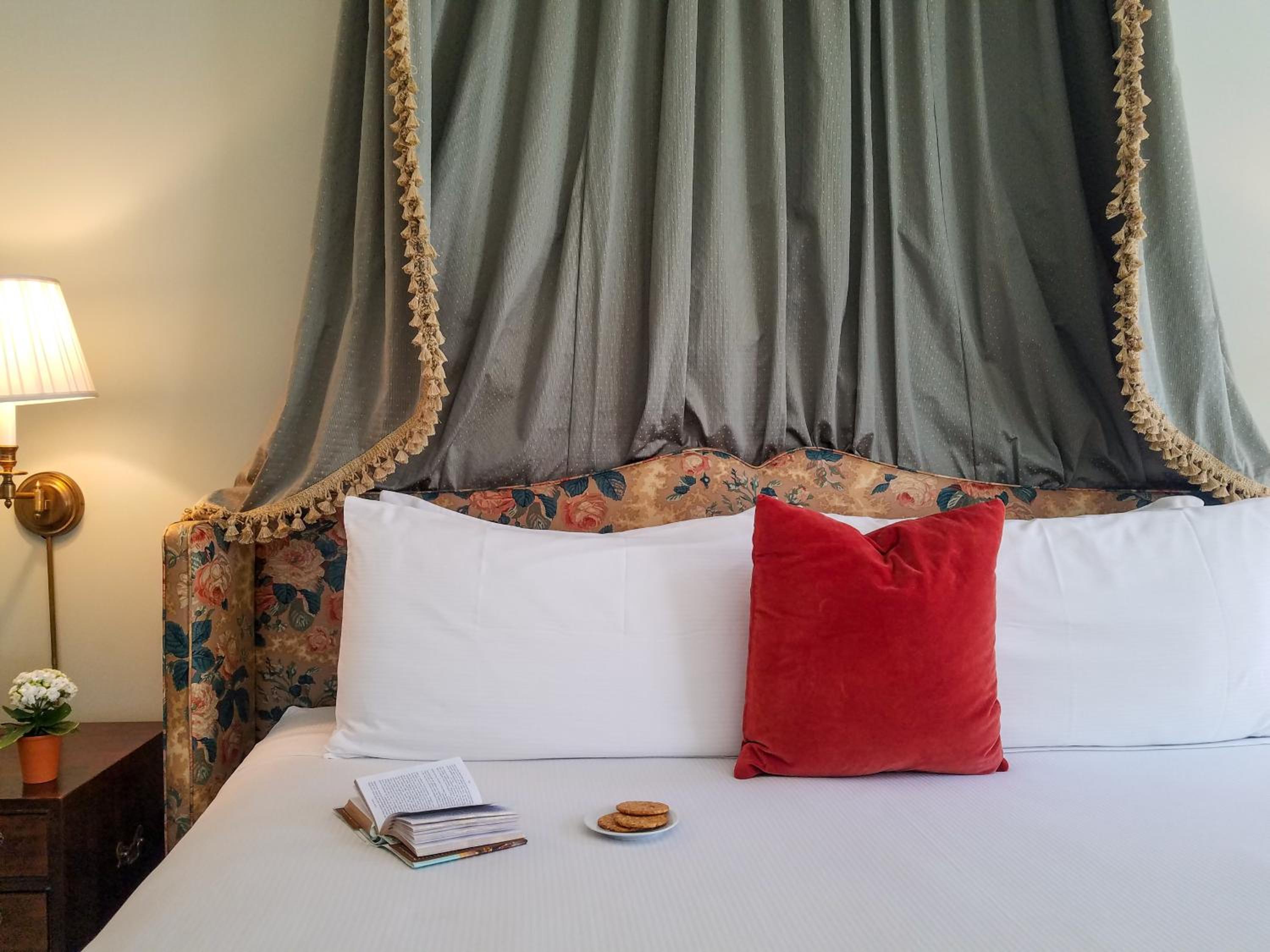A bed fitted with a fabric canopy, fringed tassel trim, floral headboard, and a bright red accent pillow.