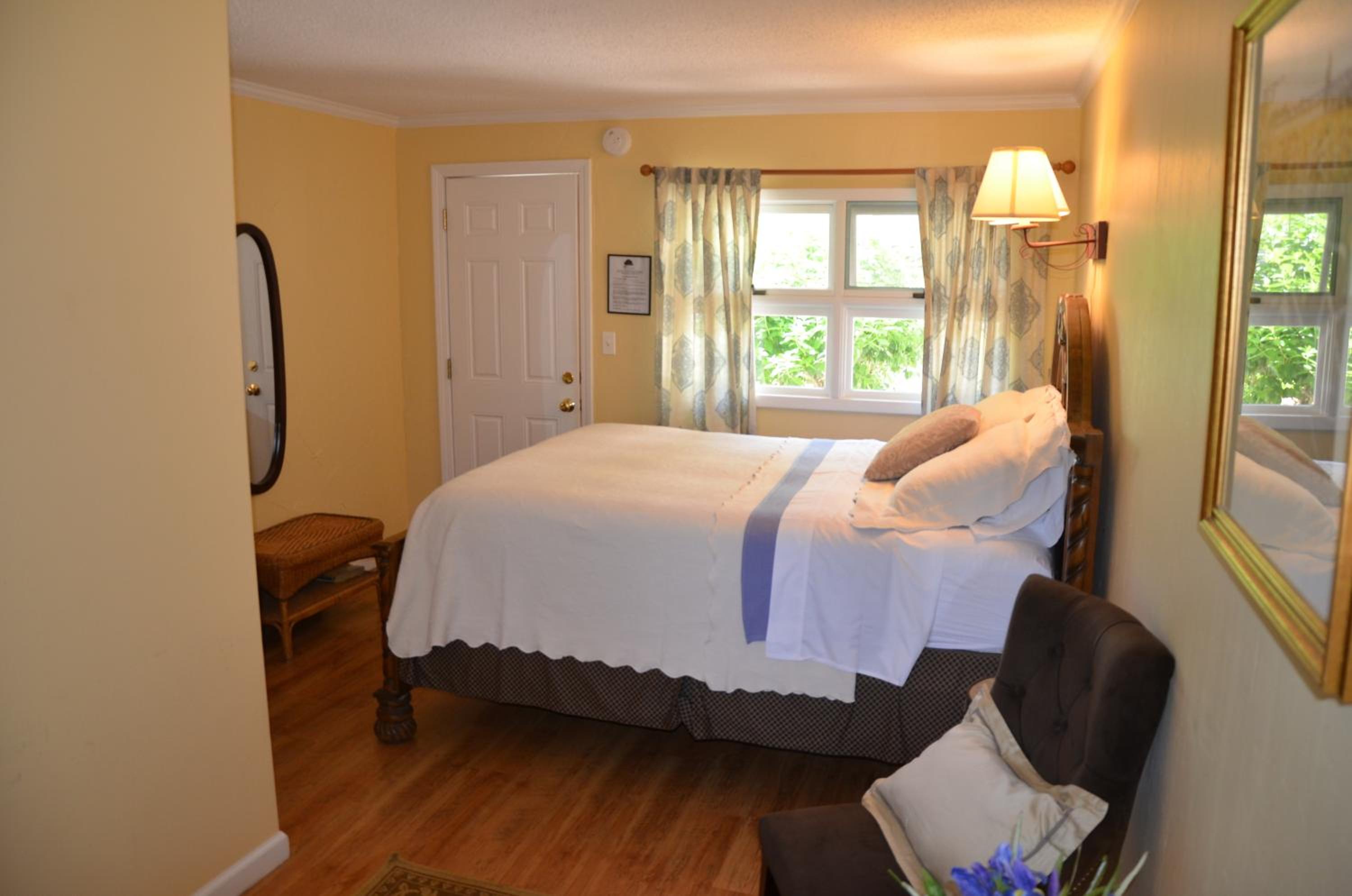 A bedroom with light yellow walls, wooden floors, an upholstered armchair, and a bed beneath a curtained window.