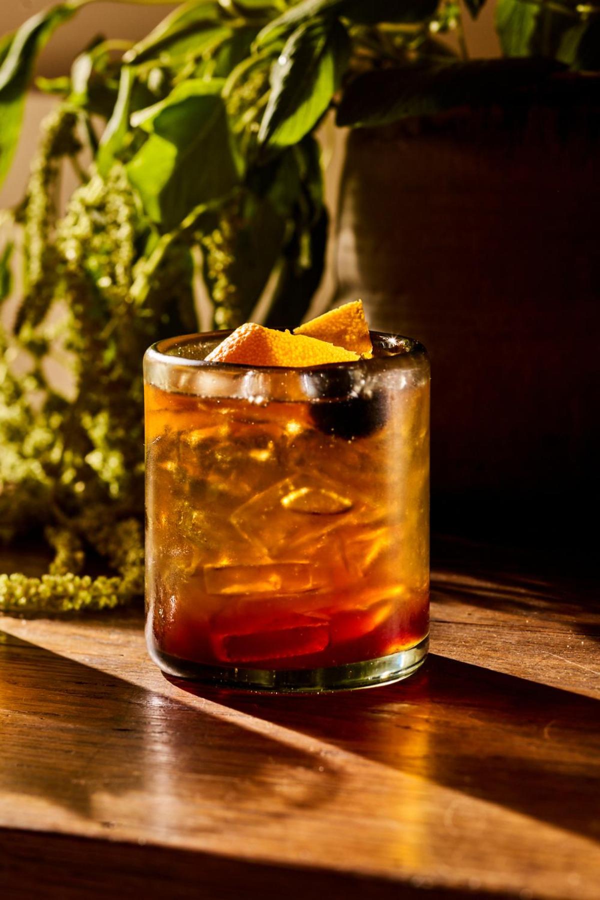 A cocktail with ice, an orange peel, and a dark cherry rests on a wooden table in sunlight.
