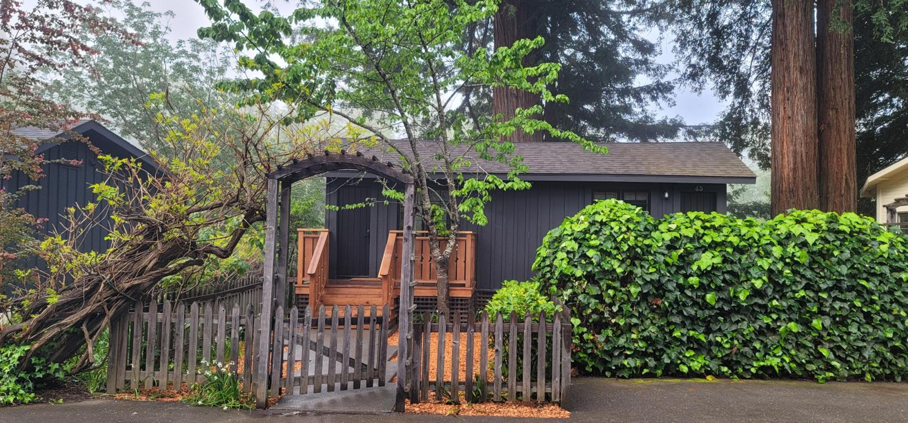 A dark cabin with a wooden picket fence and arched arbor is framed by green hedges and tall trees at Dawn Ranch.