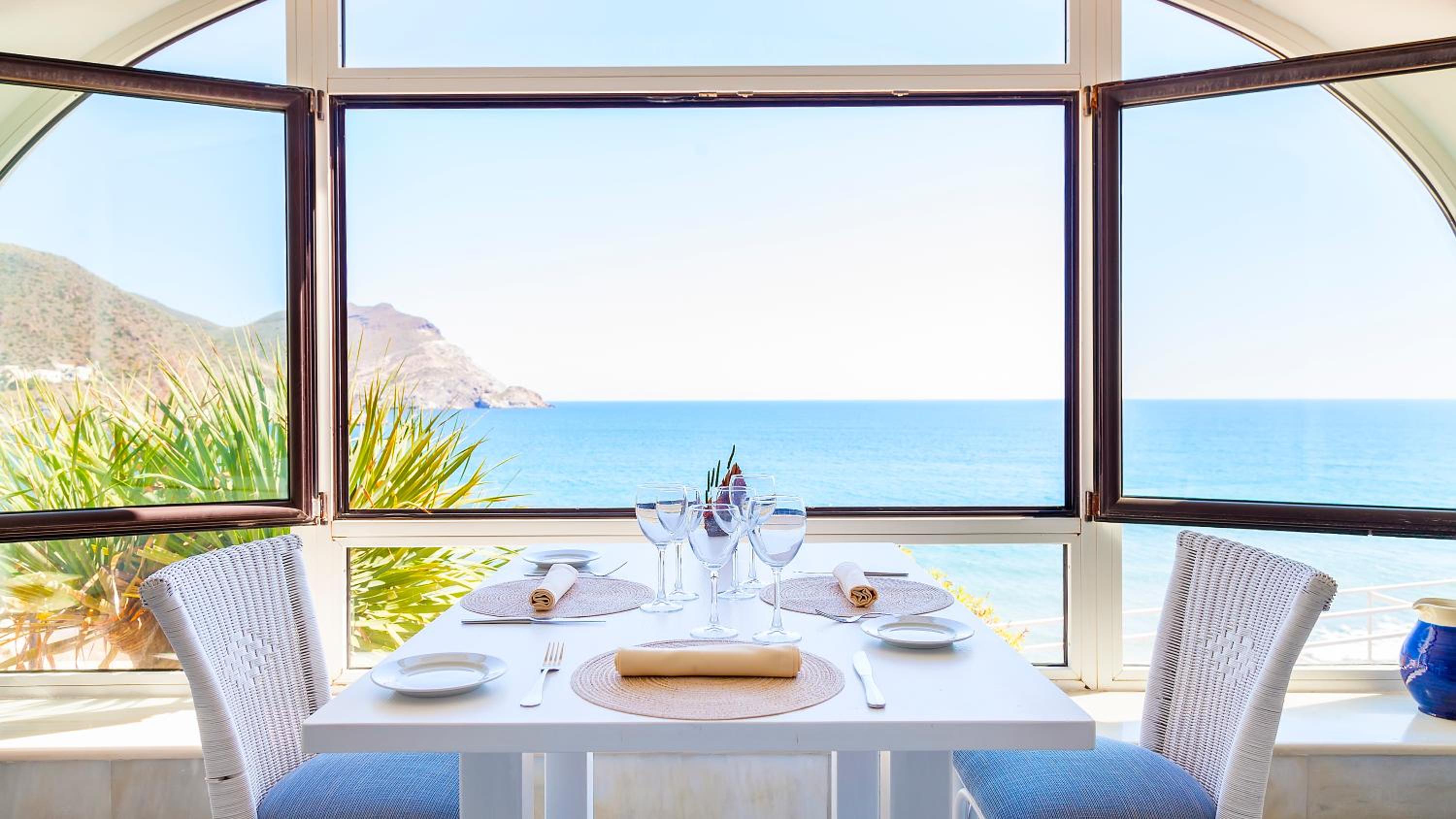 A dining table set with glassware and plates faces a wide picture window looking out to the open sea.