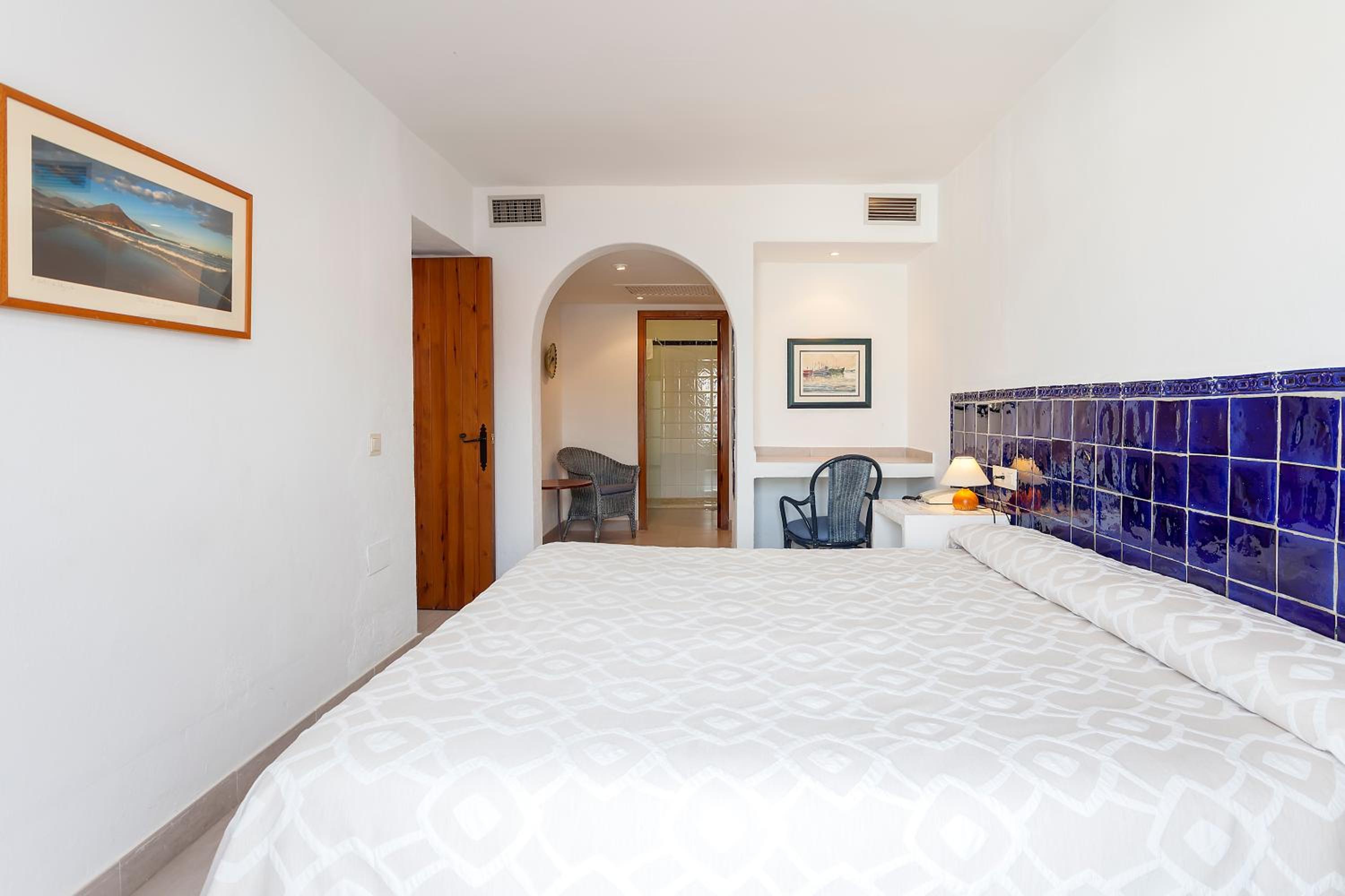 A white bedroom displays a bed with patterned covers, glossy blue headboard tiles, and an open archway.