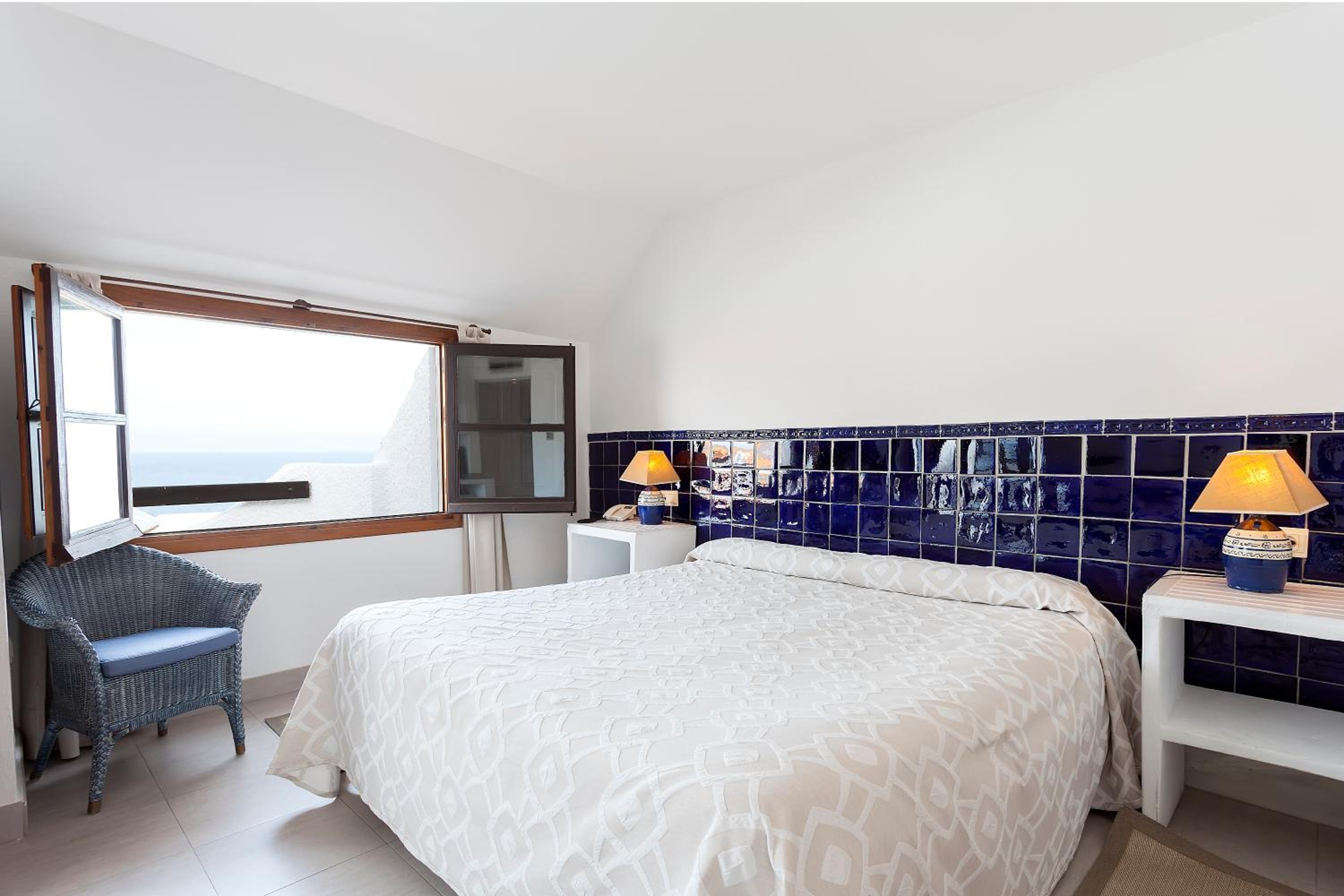 A bedroom with an open casement window offering sea views, blue wall tiles, and a woven wicker armchair.