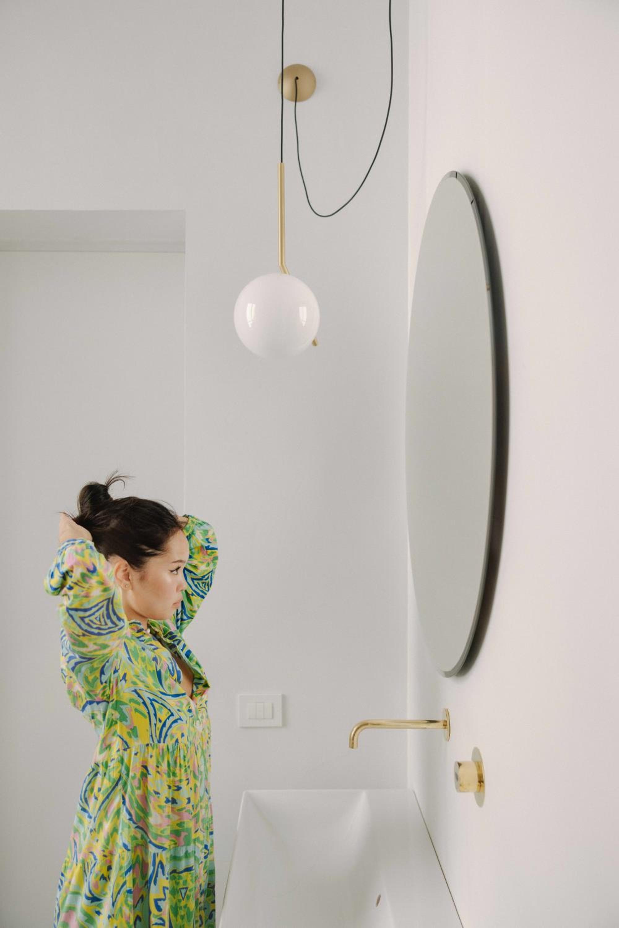 A woman adjusts her hair in front of a round mirror and brass faucet in a bright white bathroom.