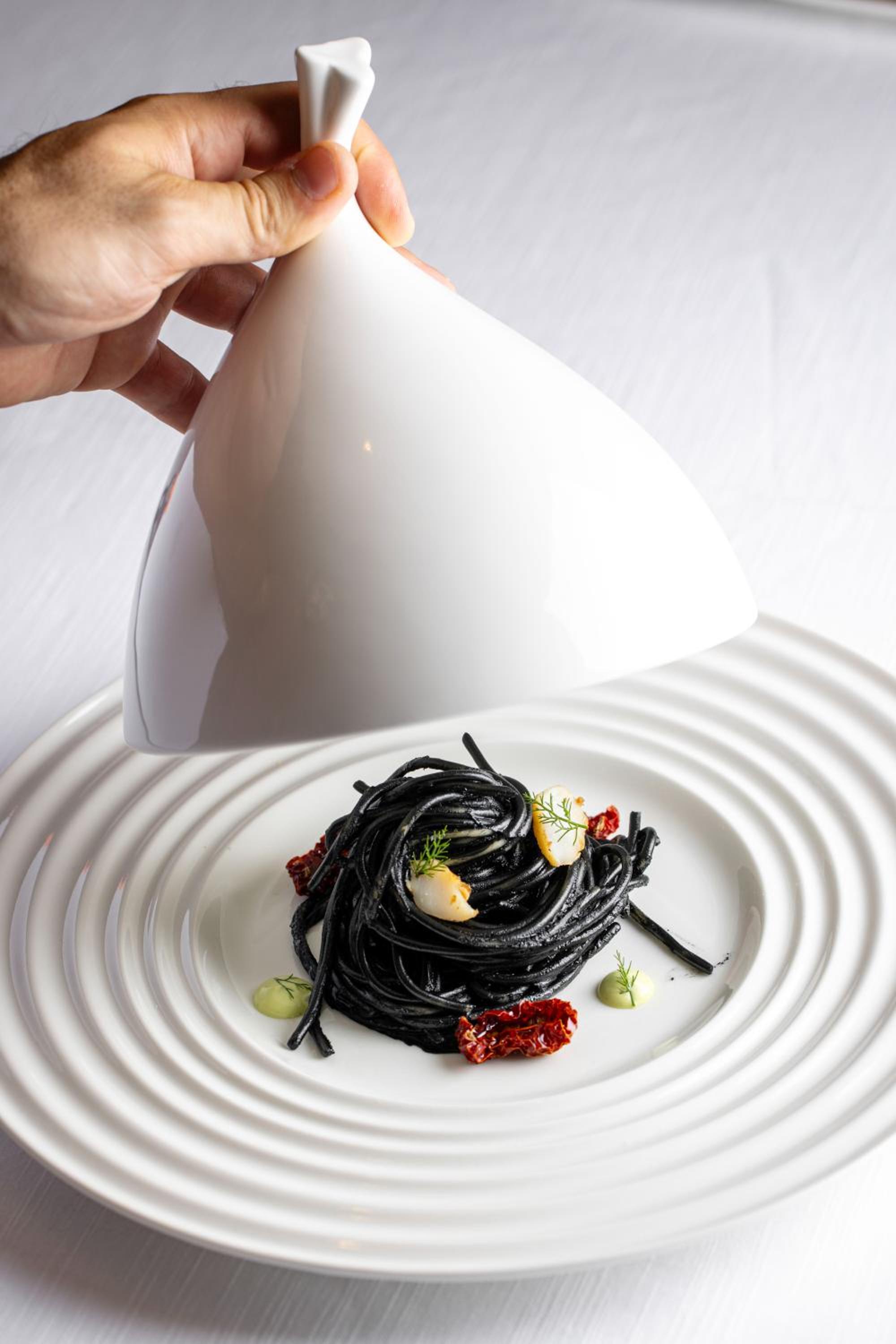 A hand lifts a white ceramic cloche over black squid-ink pasta garnished with seafood on a ridged plate.