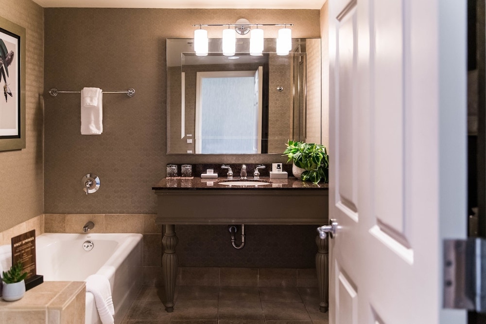 A hotel bathroom featuring a vanity with a large mirror, overhead lighting, a bathtub, and tiled flooring.