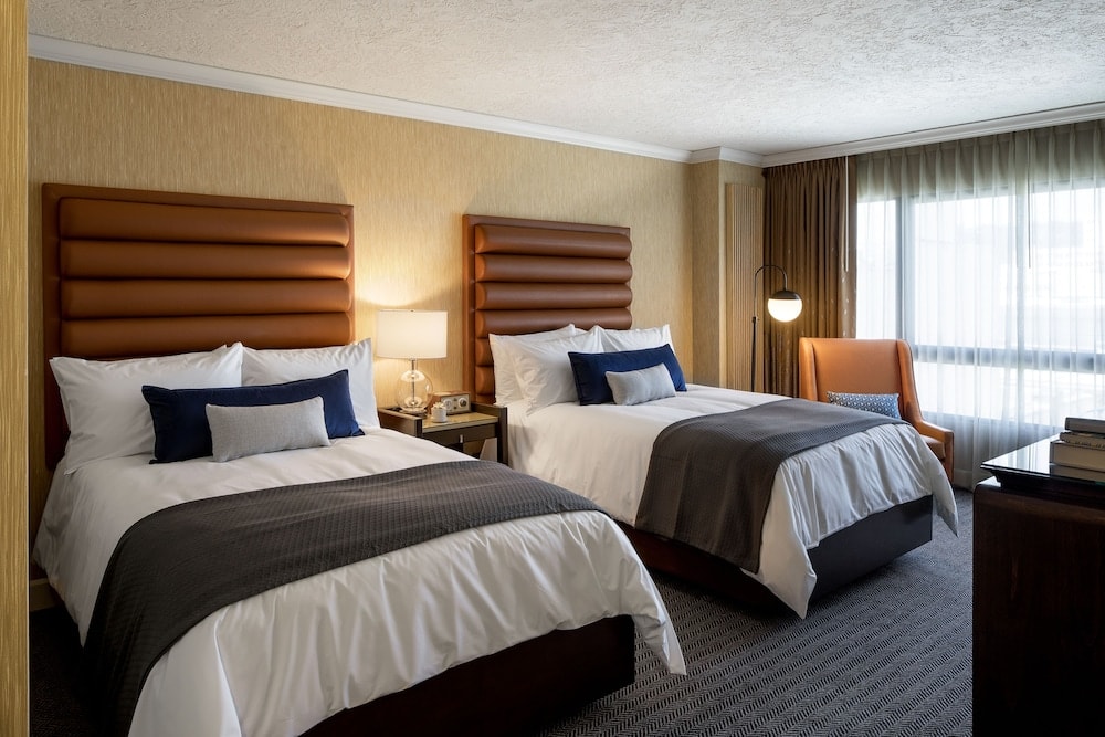 A guest room with ribbed leather headboards on the beds, white linens, an armchair, and a window with sheer curtains.