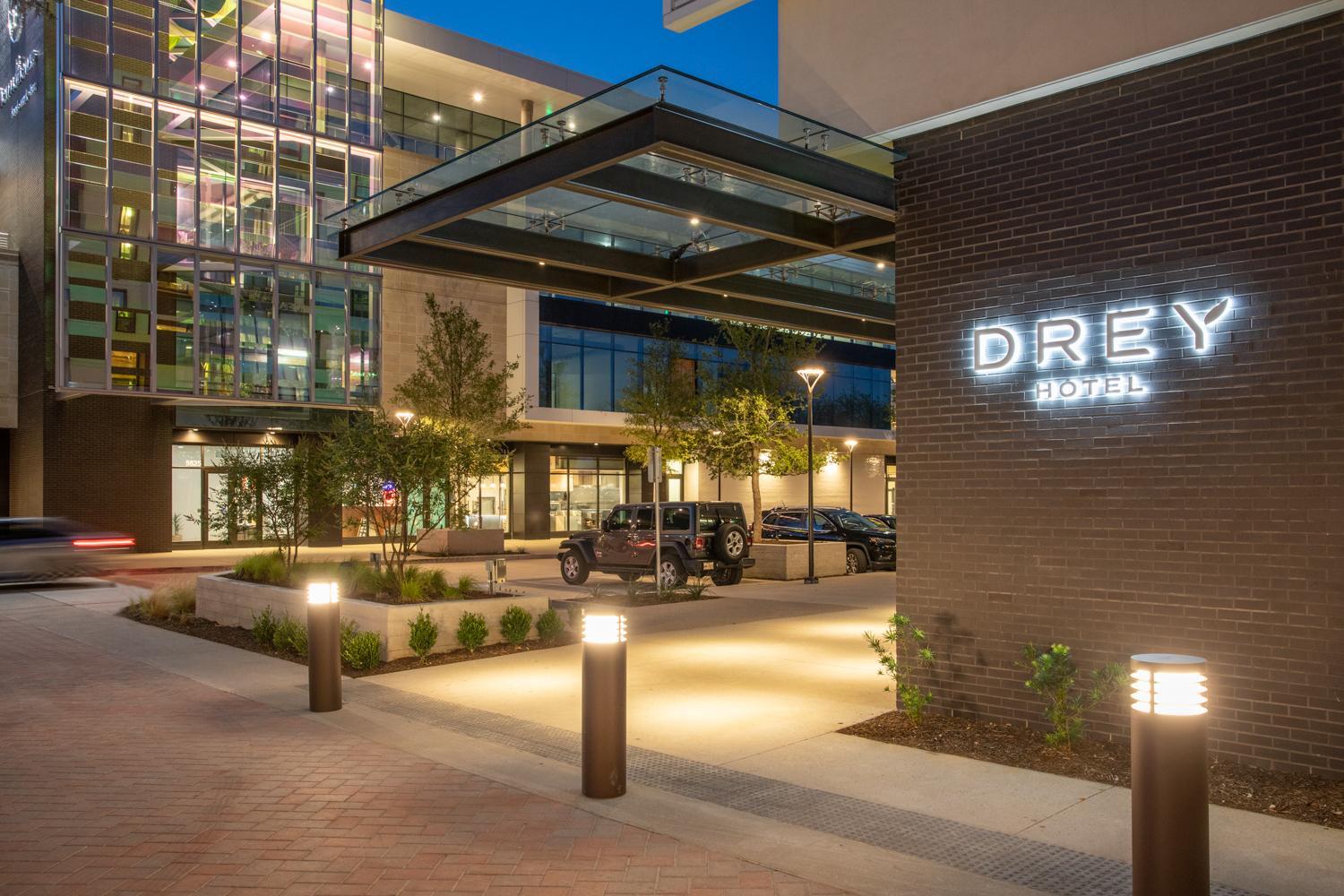 Exterior brick facade of Drey Hotel at dusk with illuminated signage and a glass canopy over the paved entryway.
