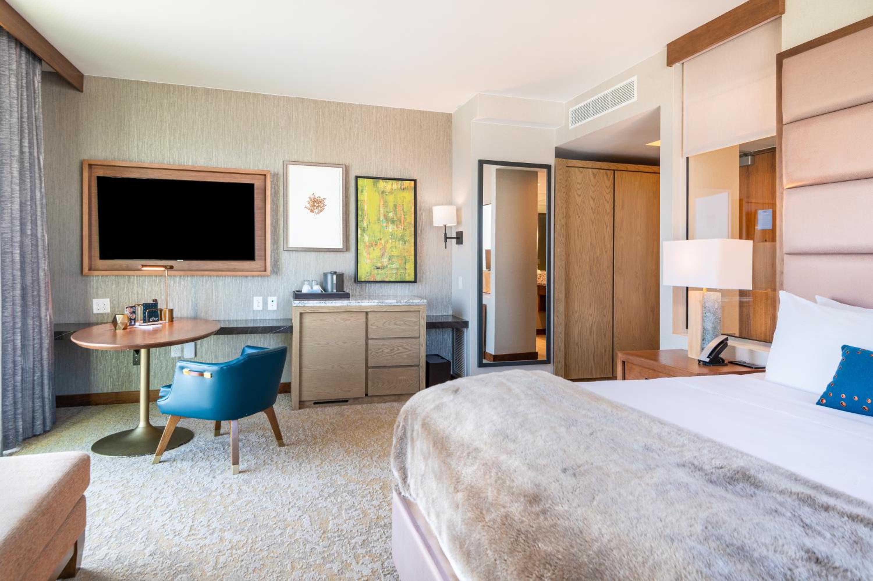 Hotel bedroom with a wall-mounted TV, round wooden desk, teal chair, and a bed with a faux-fur throw.