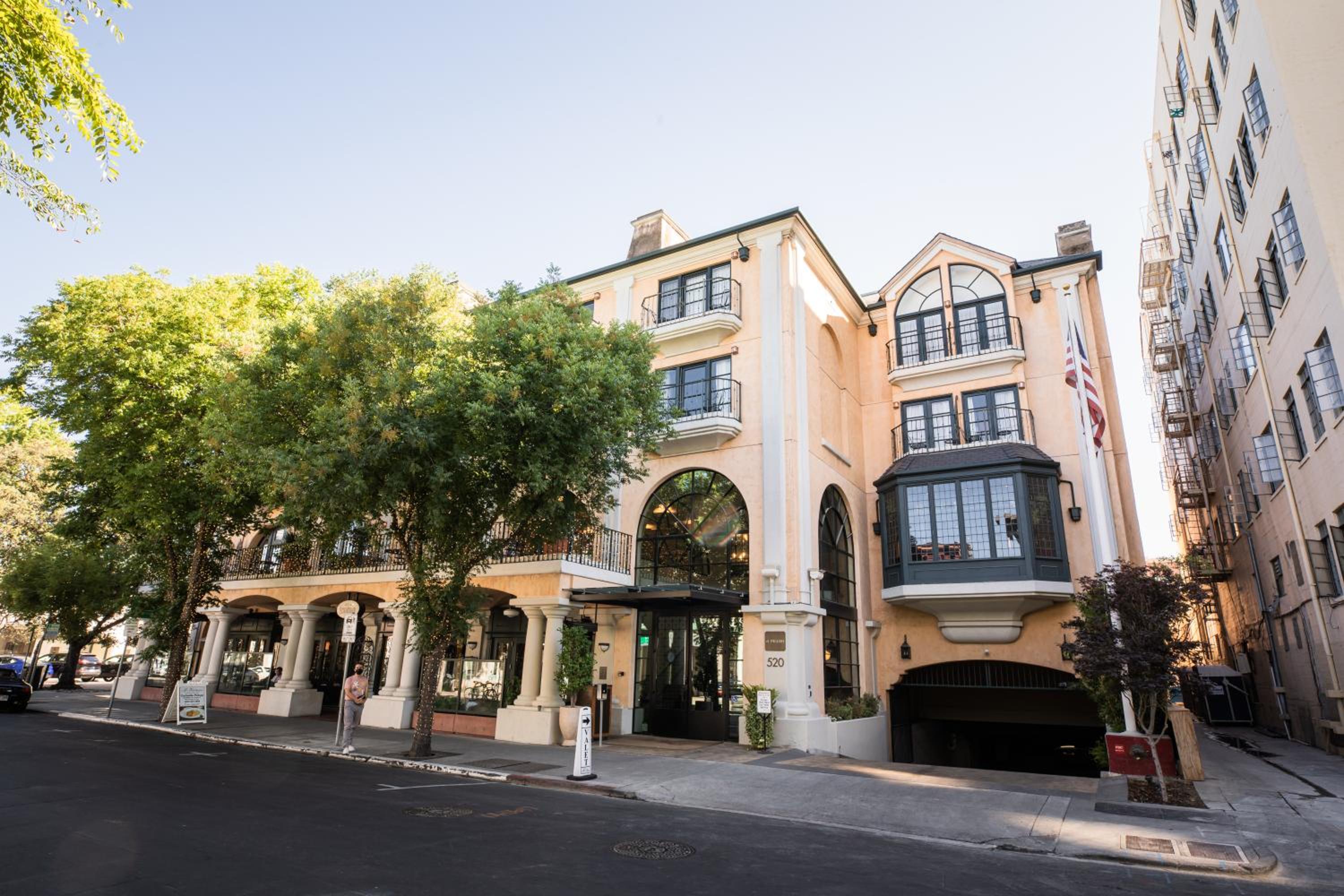 Facade of el PRADO hotel featuring arched windows, stone pillars, upper balconies, and leafy trees along the street.