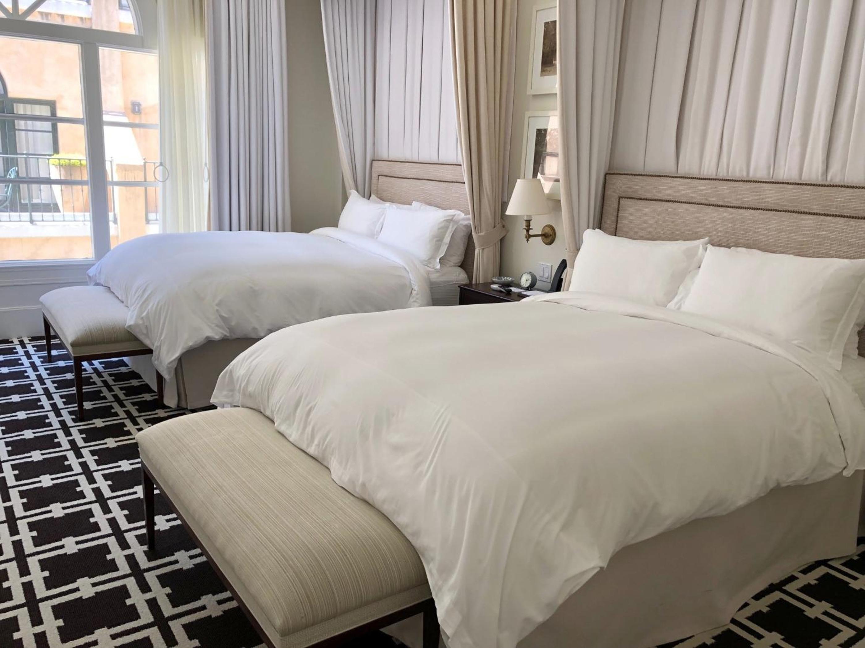 Guest room with beds dressed in white linens, upholstered headboards, a patterned rug, and sheer drapes by a terrace door.