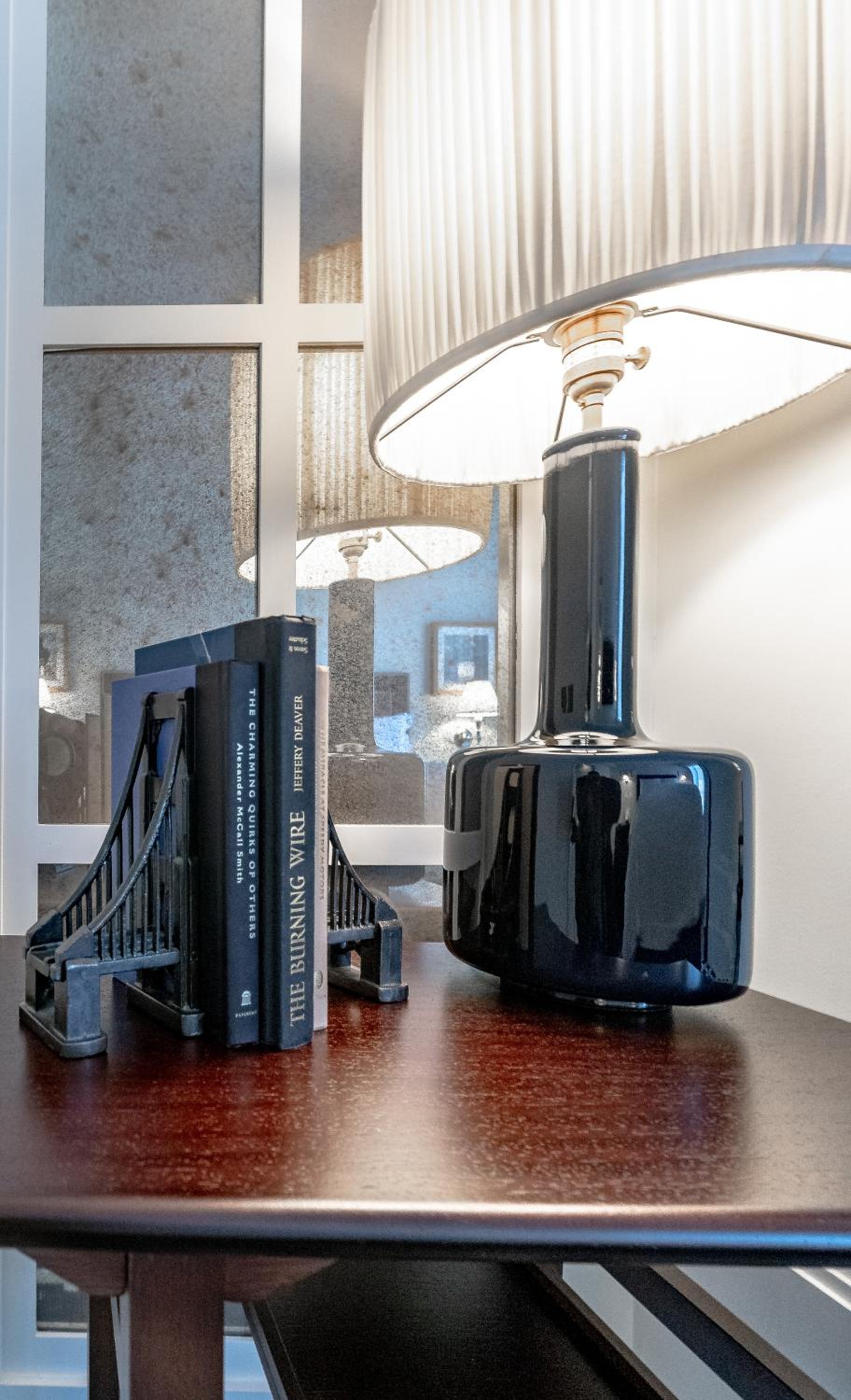 Wooden table holding books secured by bridge-shaped bookends beside a tall dark ceramic lamp with a pleated shade.