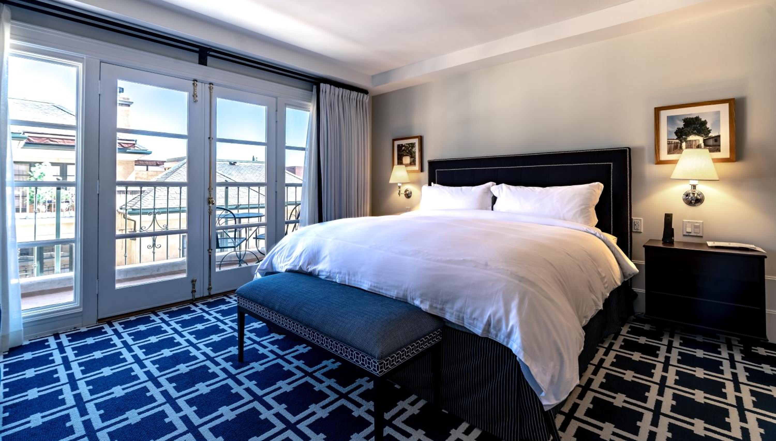 Bedroom with a dark cushioned headboard, white bedding, geometric blue carpet, and French doors opening onto a balcony.