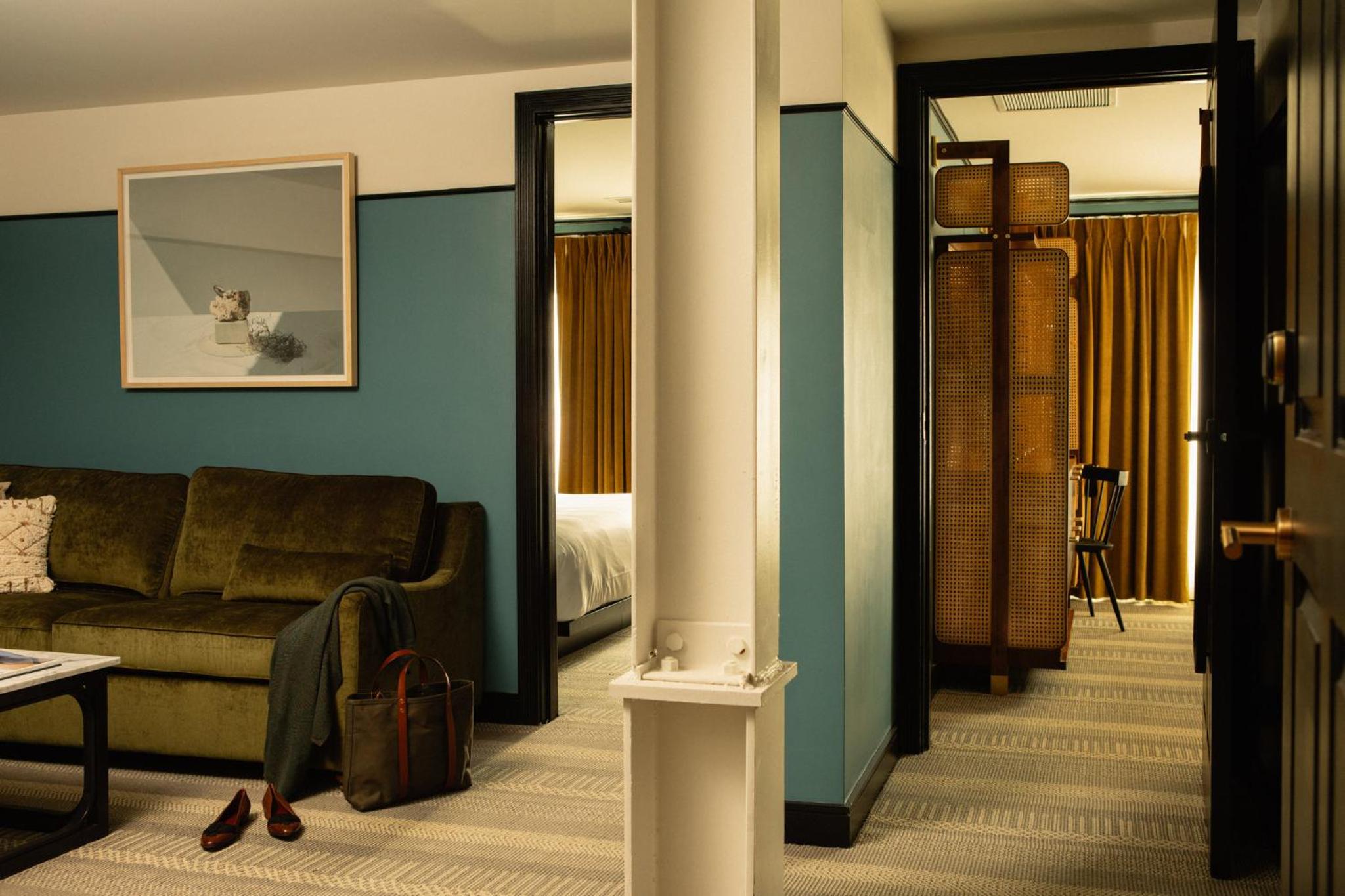 A hotel suite living room with a green velvet sofa and teal walls opening into an adjoining bedroom.