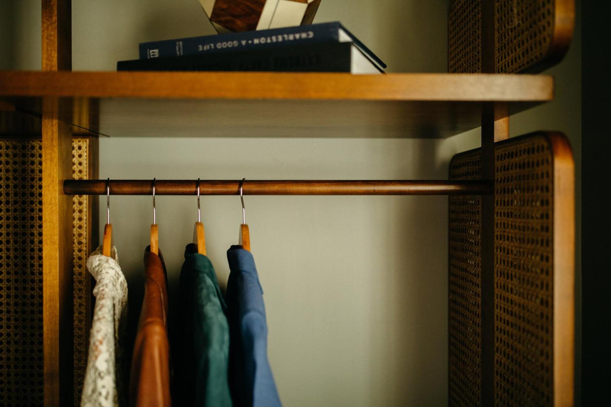 Clothes hang on wooden hangers inside a cane and timber wardrobe beneath a shelf holding books.