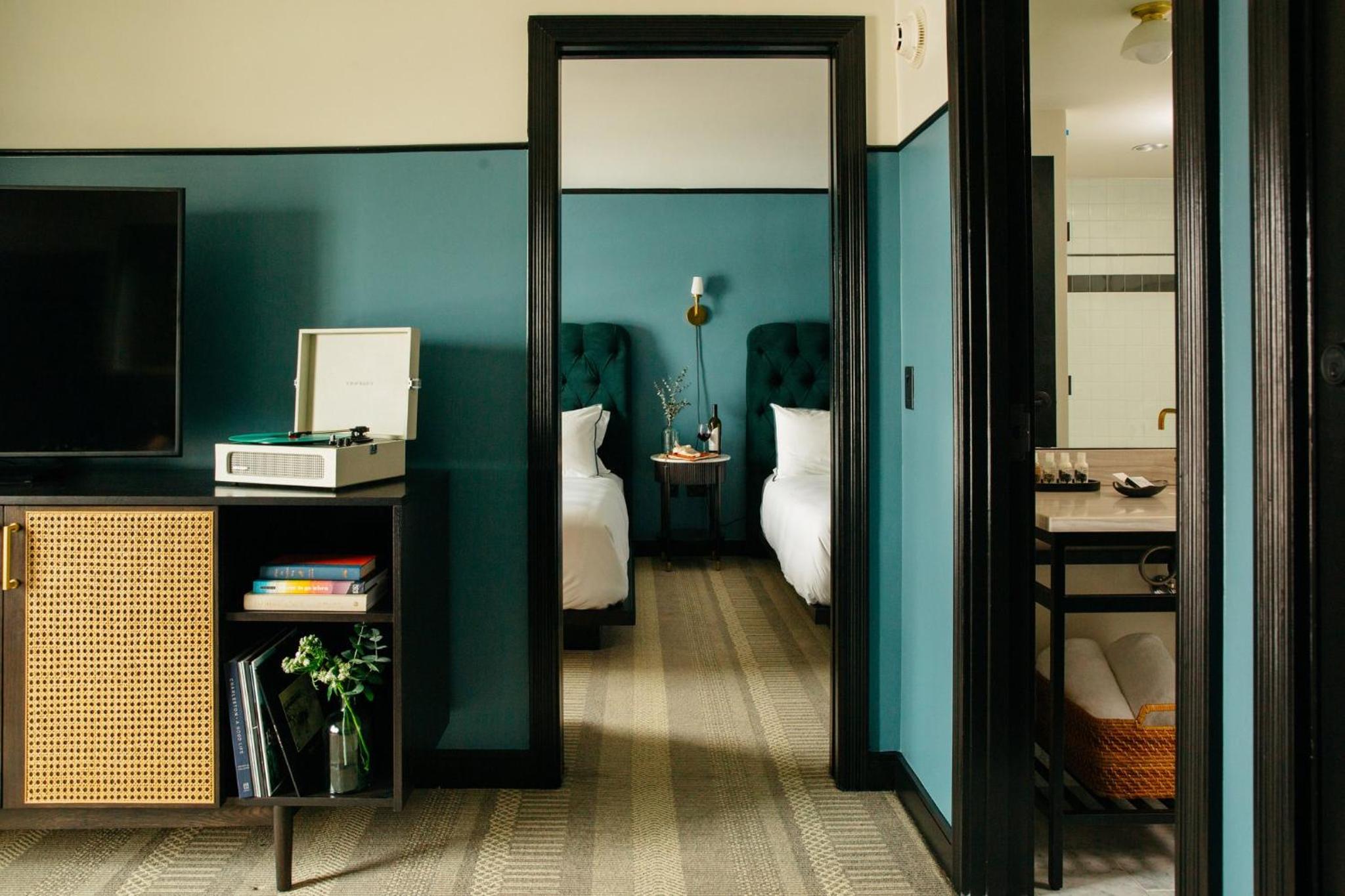 A credenza with a record player stands beside a doorway leading into a bedroom with teal velvet headboards.