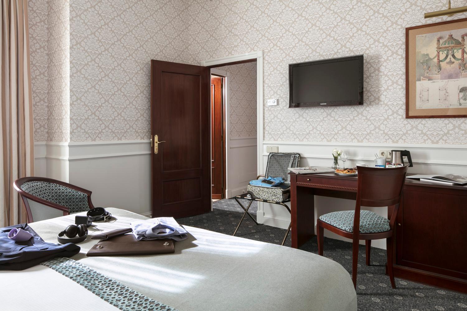 A hotel bedroom with patterned wallpaper, a dark wood writing desk, and a television mounted on the wall.