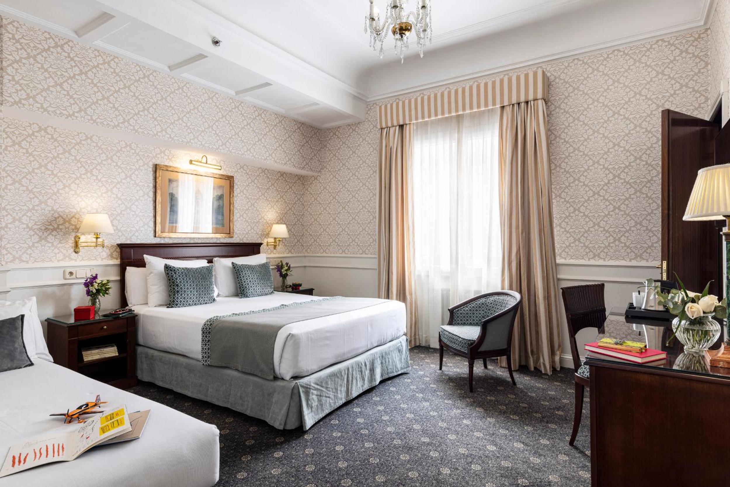 A bedroom with patterned damask wallpaper, a made bed, striped window drapes, and a crystal chandelier.