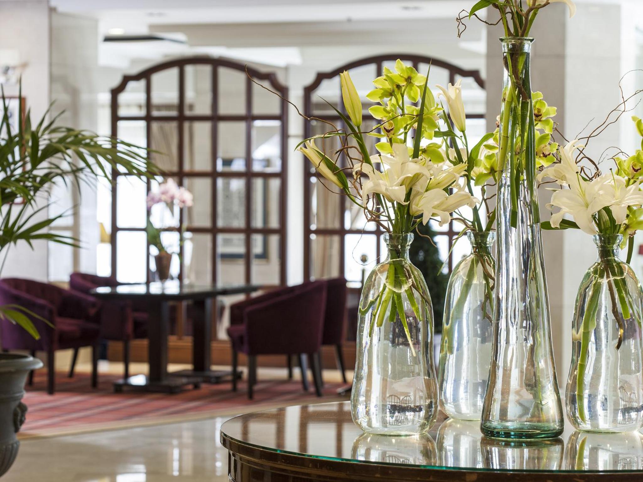Tall glass vases with white and green flowers set atop a round glass table in a lounge area.