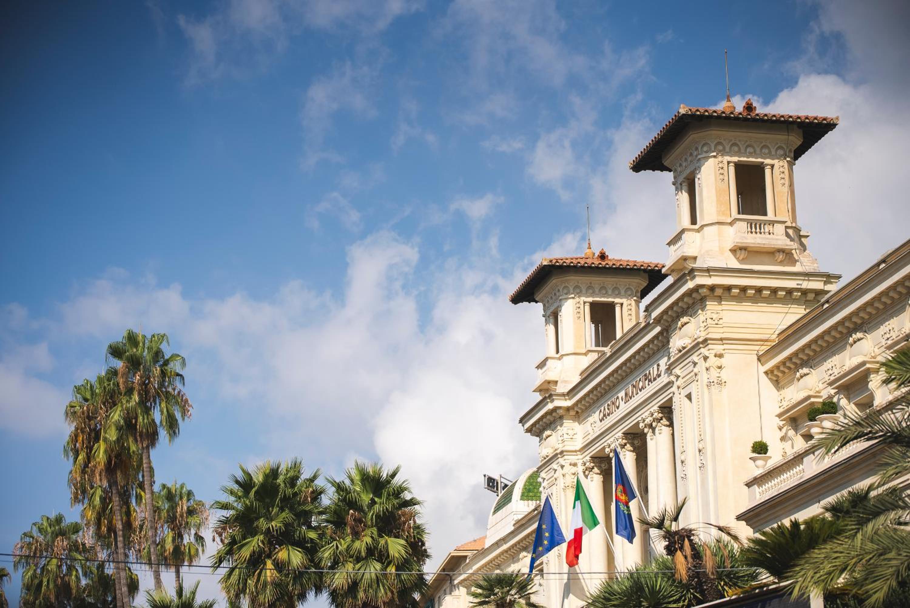 An ornate historic facade with towers, columns, and flags framed by palm trees against a blue sky.