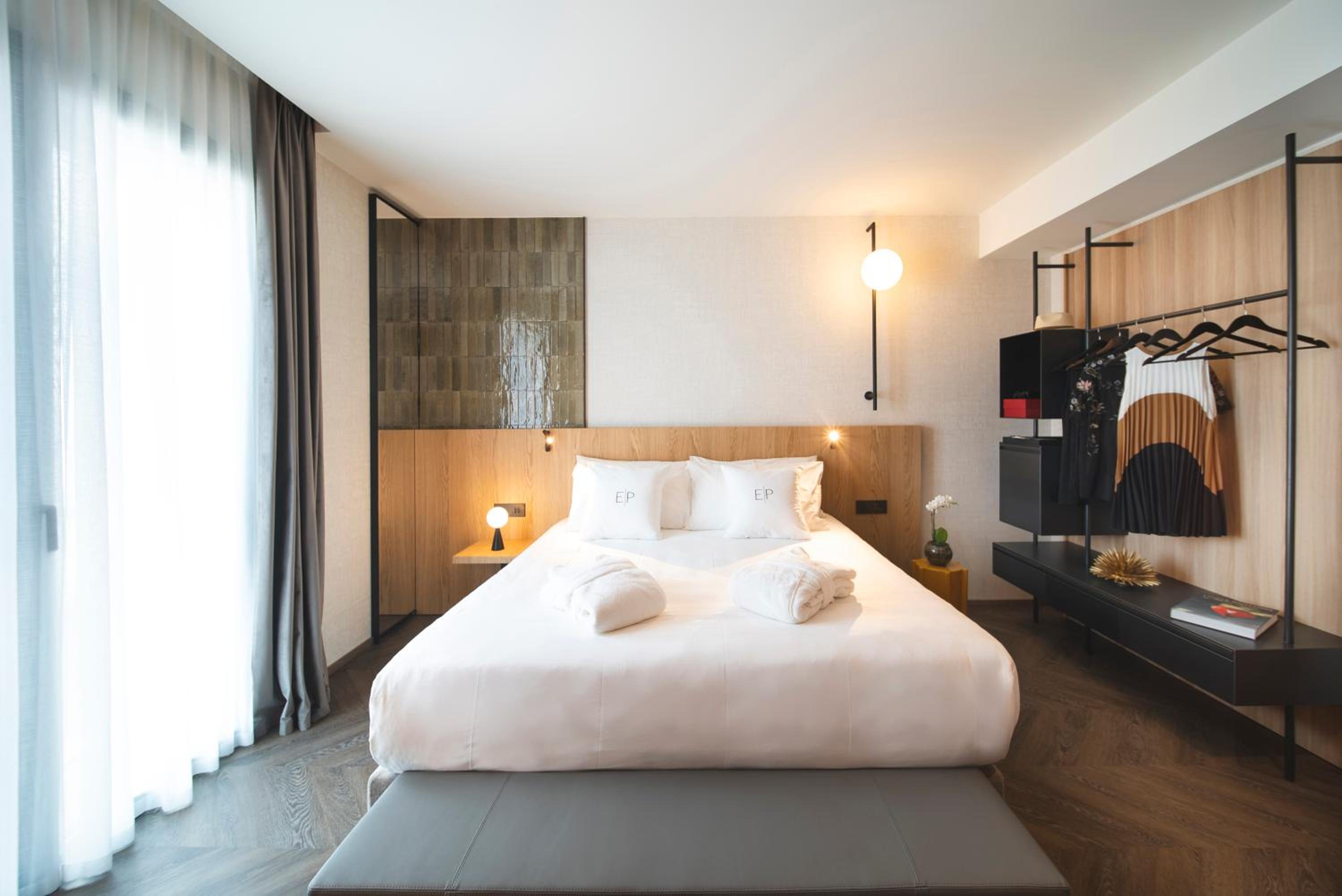 A hotel bedroom with a wood-accented wall, sheer balcony curtains, and an open wardrobe rack.