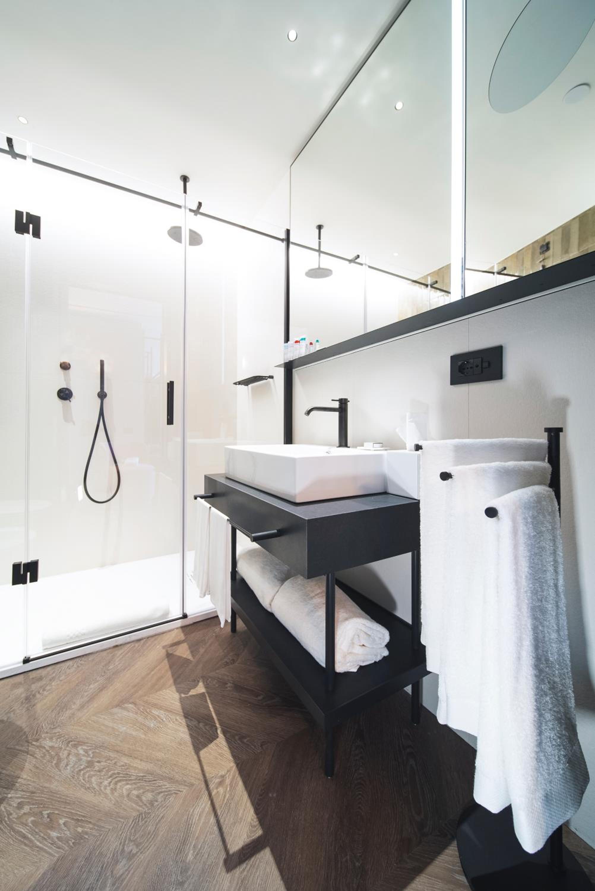 A bathroom with chevron wood flooring, a glass walk-in shower, and a black vanity with white towels.
