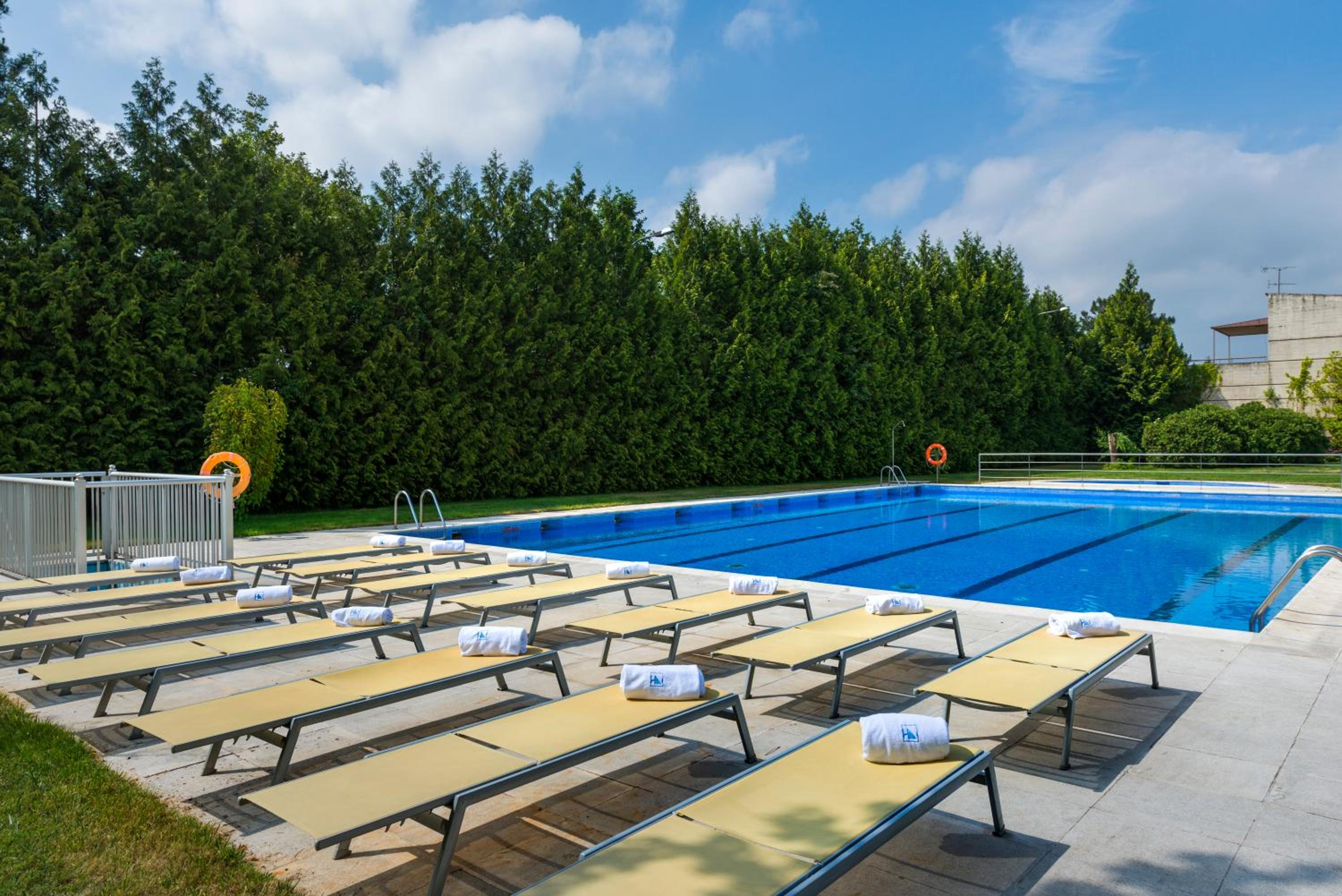 An outdoor swimming pool bordered by sun loungers with folded towels, flanked by a tall evergreen hedge.