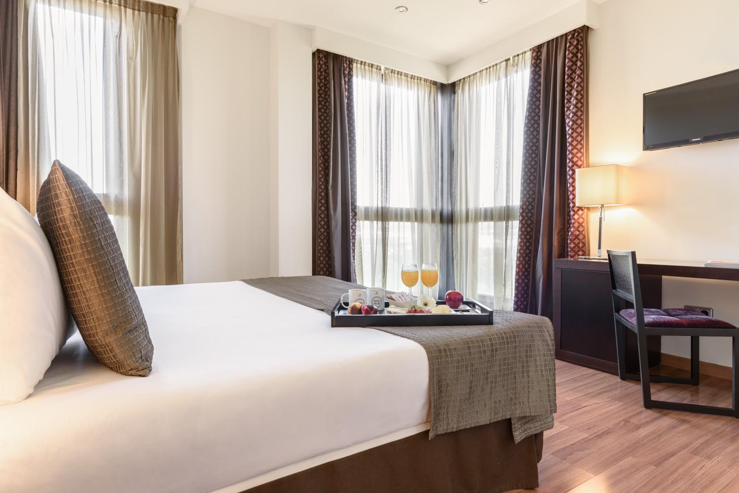 A hotel bedroom with a breakfast tray on the bed, corner windows draped with sheer curtains, and a desk.