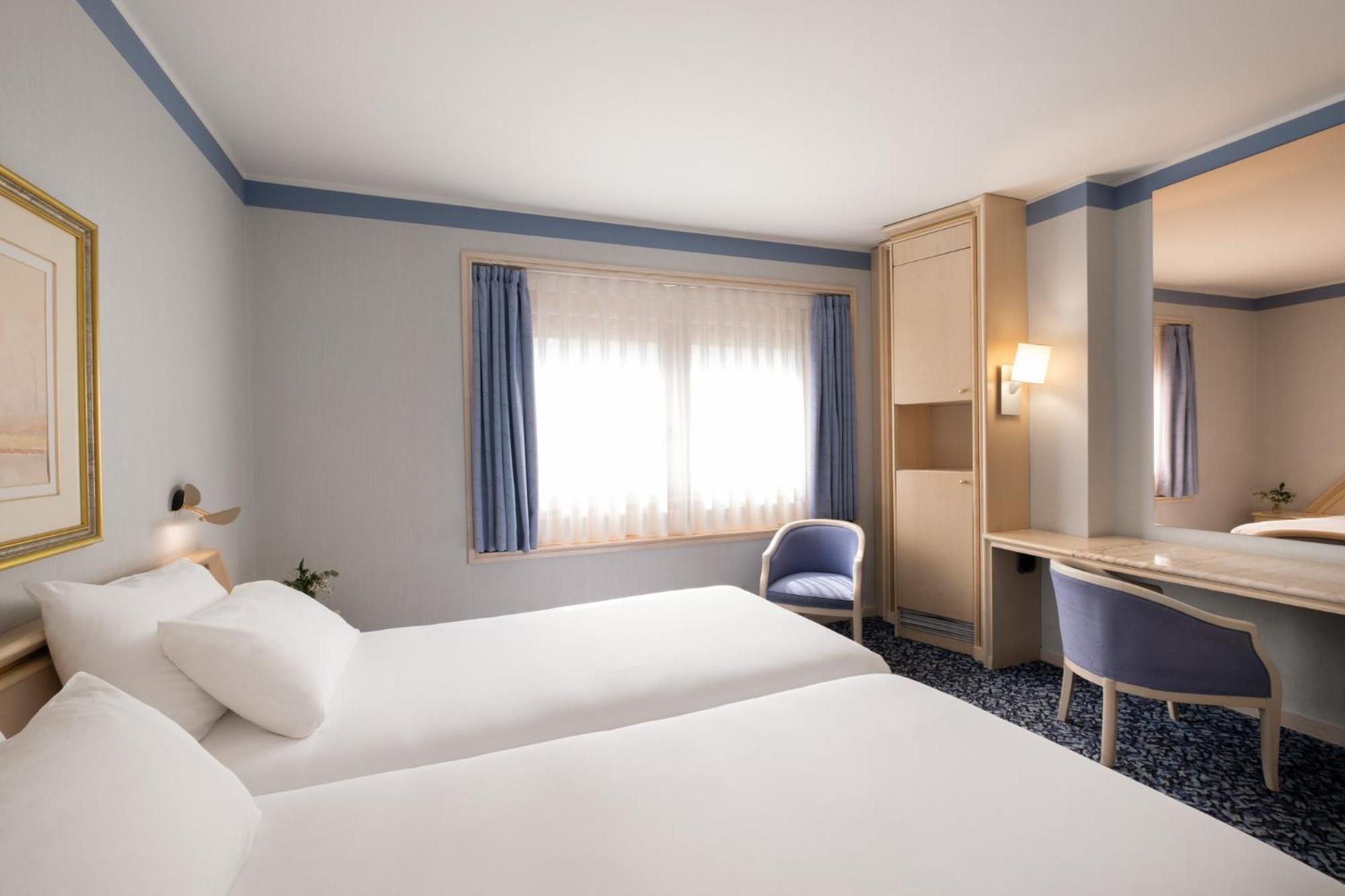 Hotel bedroom at Exe Boston with white beds, blue armchairs, patterned carpeting, and a desk.