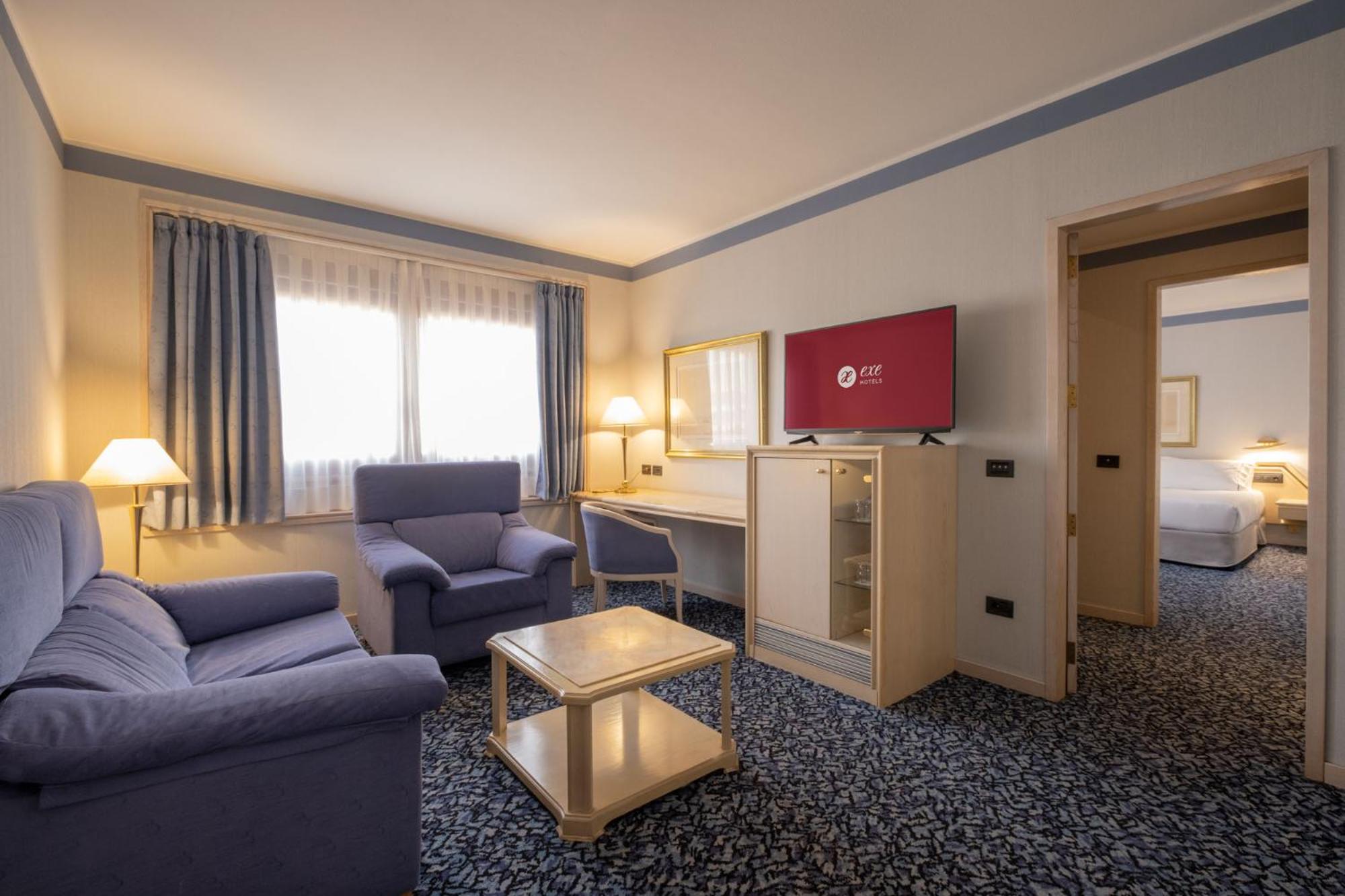 Hotel sitting room with blue upholstered seating, a coffee table, patterned carpet, and a doorway to a bedroom.