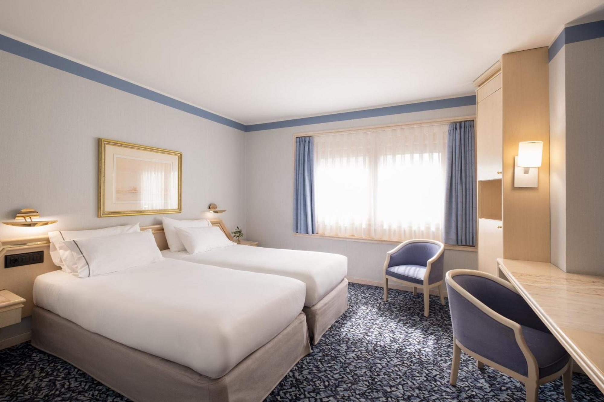 Hotel bedroom with white bedding, patterned carpet, a writing desk, and blue curtains over a window.