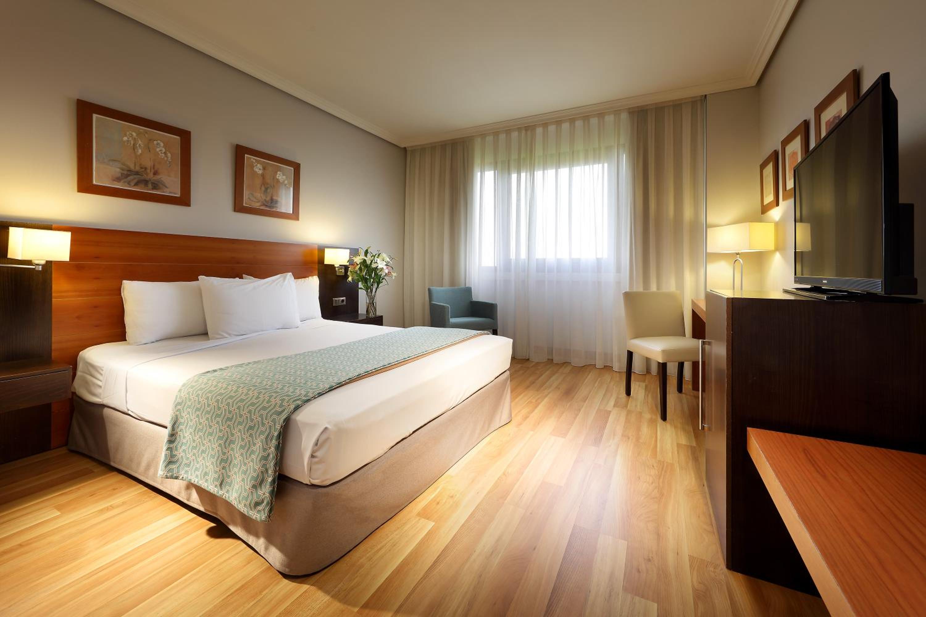 A hotel room at Exe Getafe with a large made bed, wooden headboard, light wood floors, and sheer window drapes.