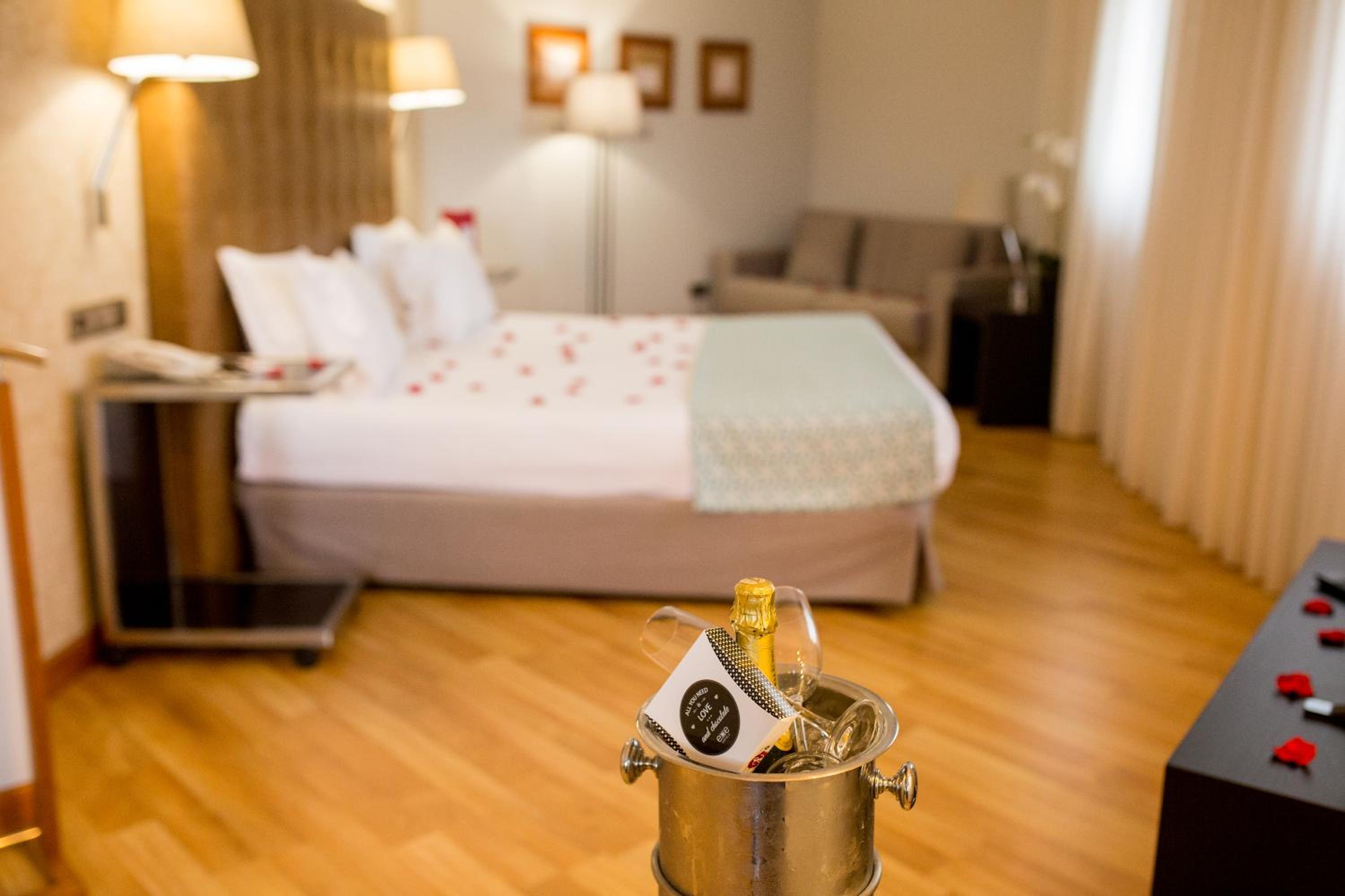 A wine cooler with glasses stands in the foreground before a hotel bed sprinkled with rose petals.