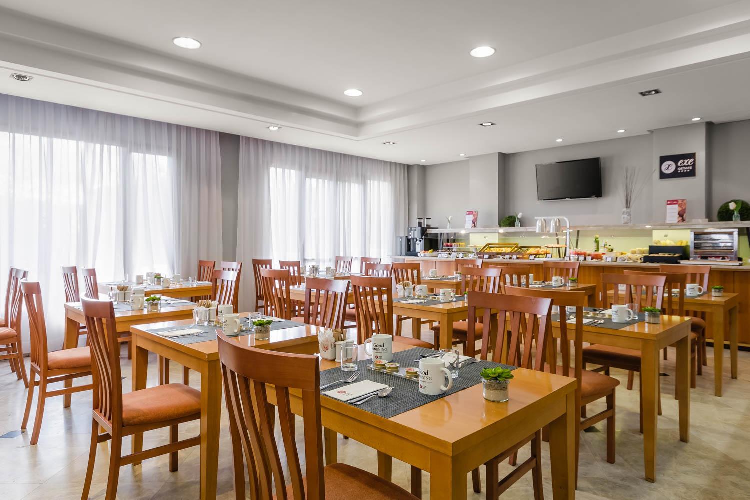 A bright dining room with wooden tables, chairs, place settings, and a food buffet along the back wall.