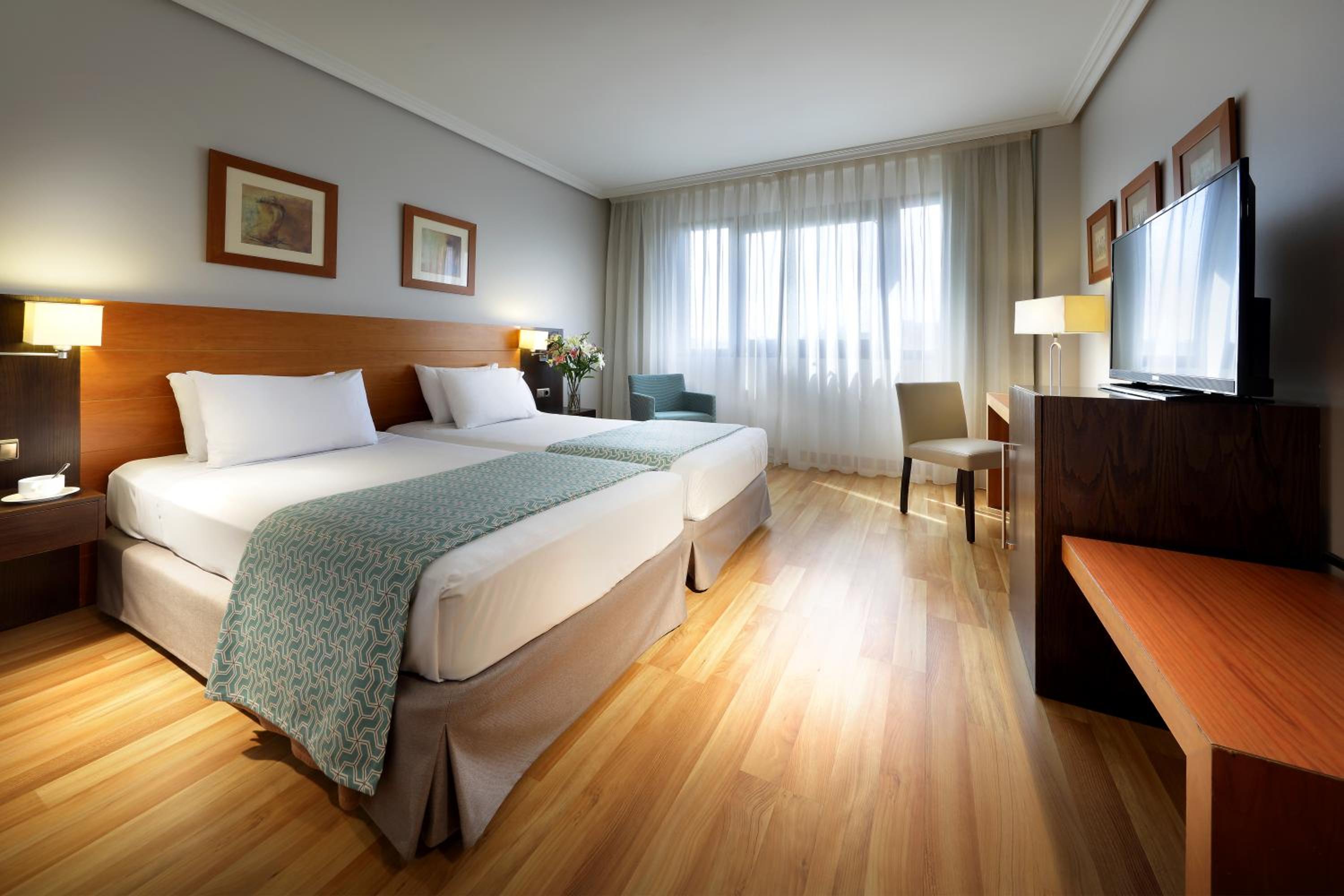A hotel bedroom with made beds, light wood flooring, a television desk, and sheer curtains over a window.