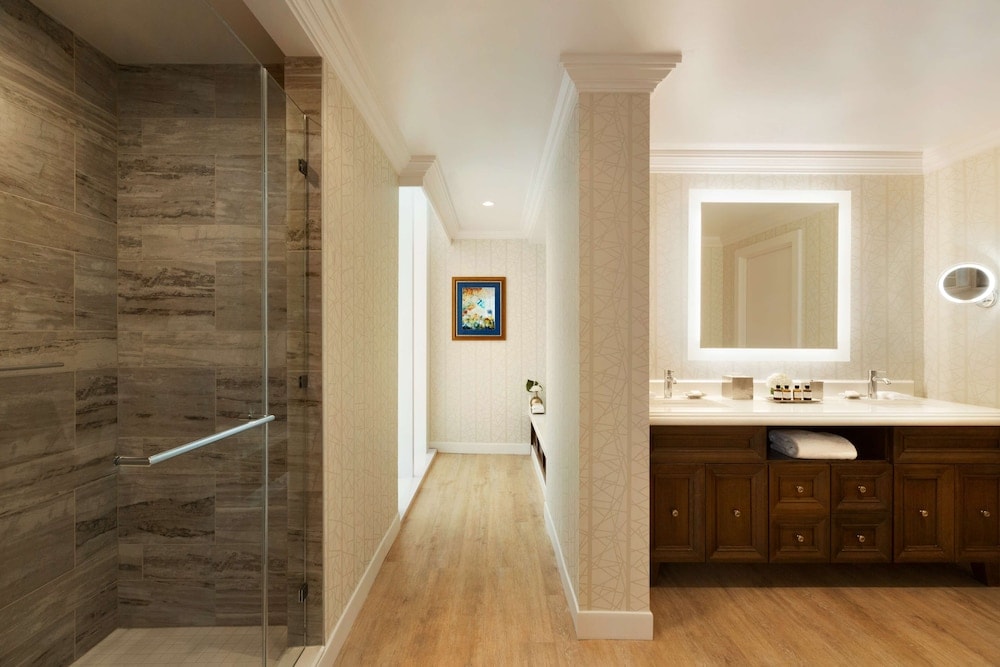 A hotel bathroom featuring a glass shower stall, wood vanity with a backlit square mirror, and light flooring.