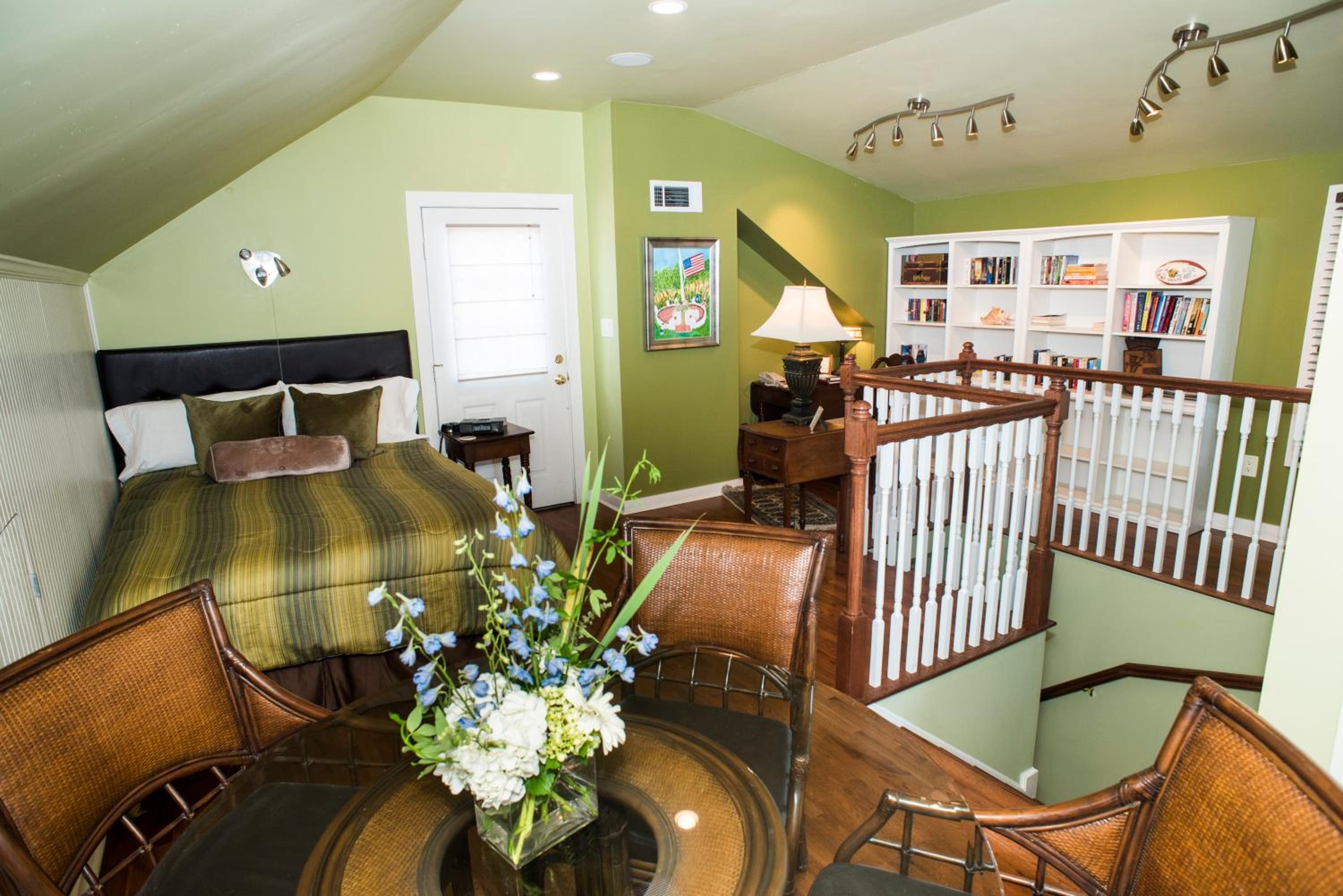 A loft bedroom with green sloped walls, a wooden railing, white built-in bookcases, and a rattan table set.