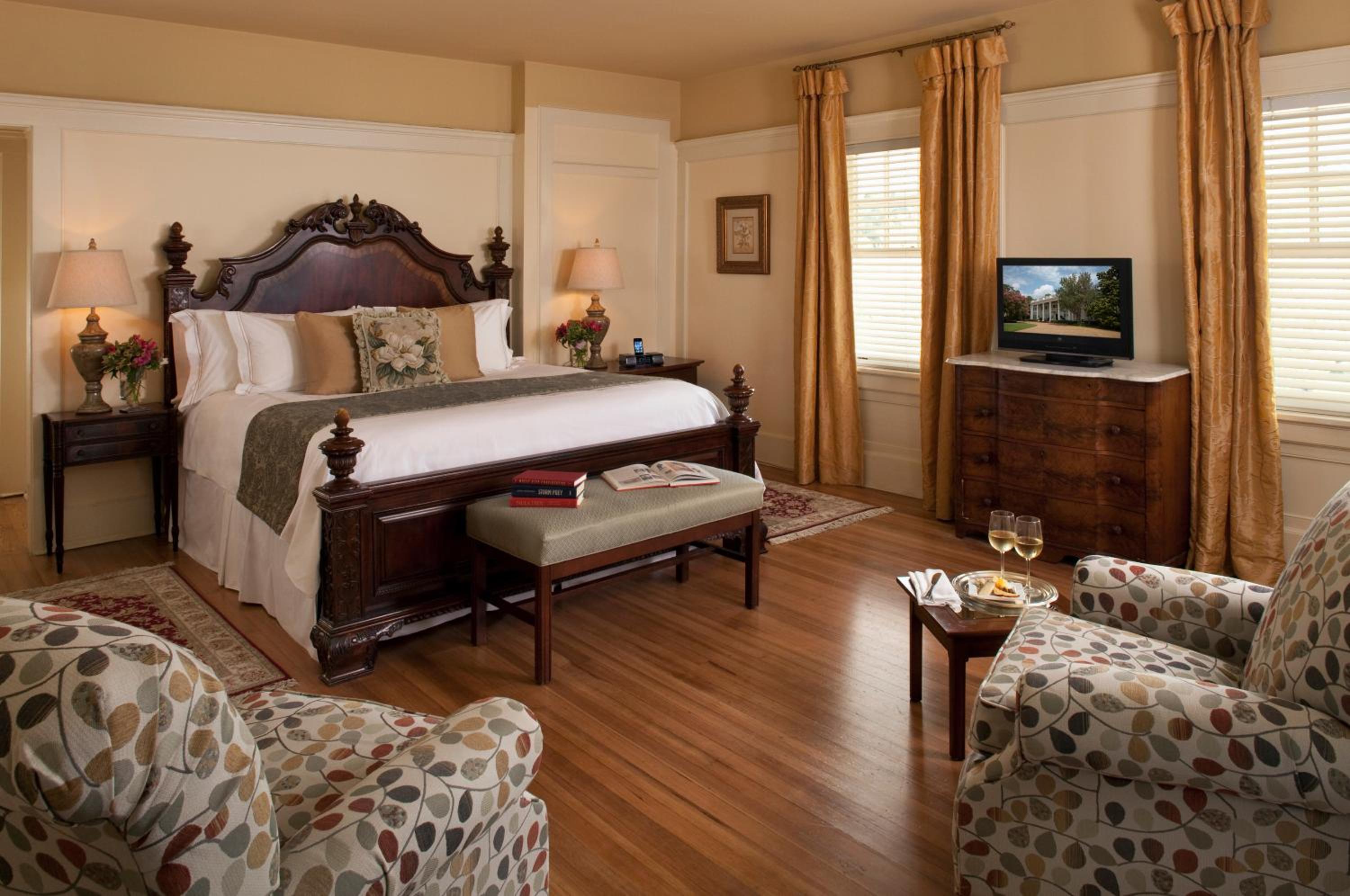 A bedroom with a dark carved wood bed, hardwood flooring, patterned armchairs, and golden drapery.