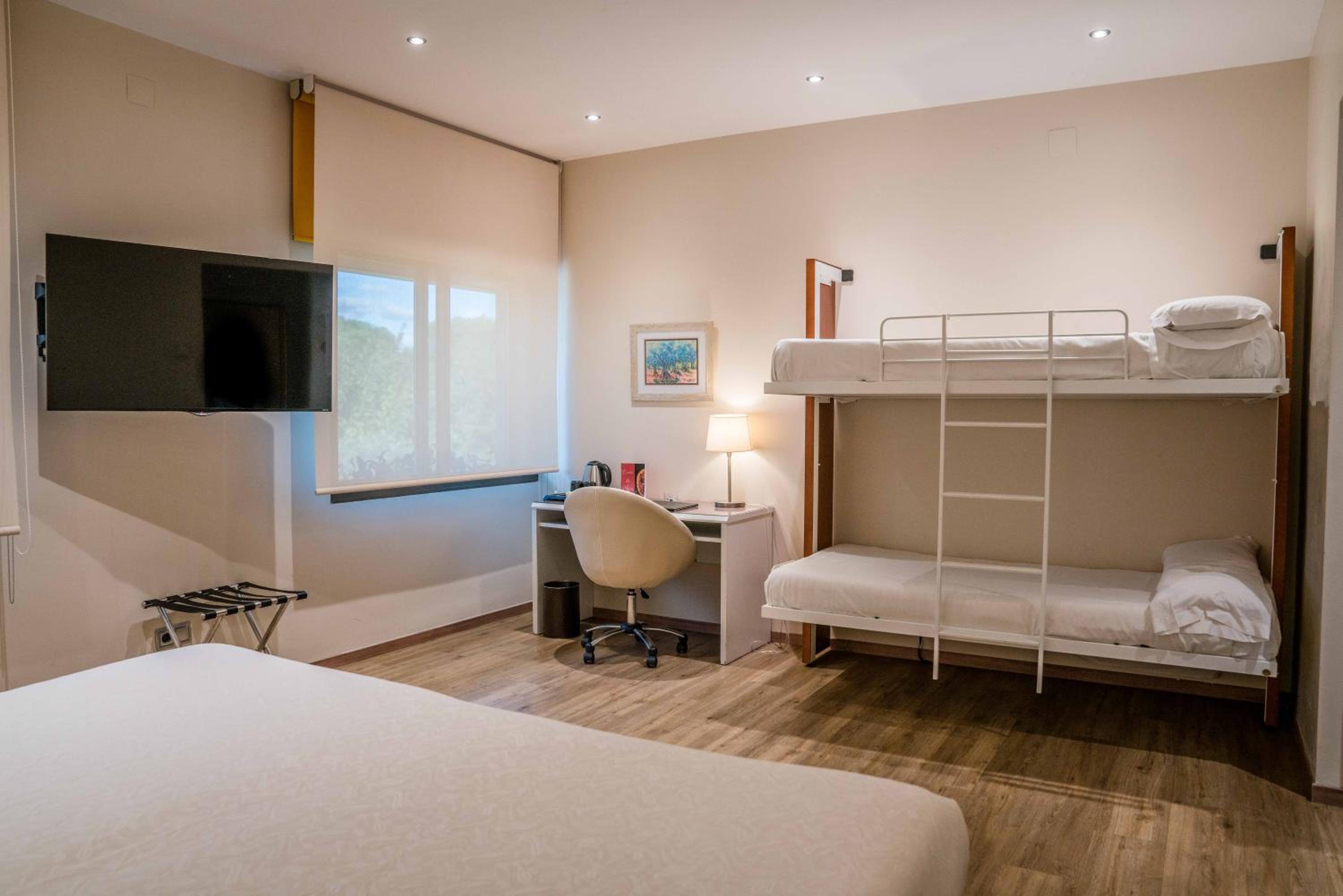 A bedroom with a work desk, wall-mounted television, and metal bunk beds against neutral walls.