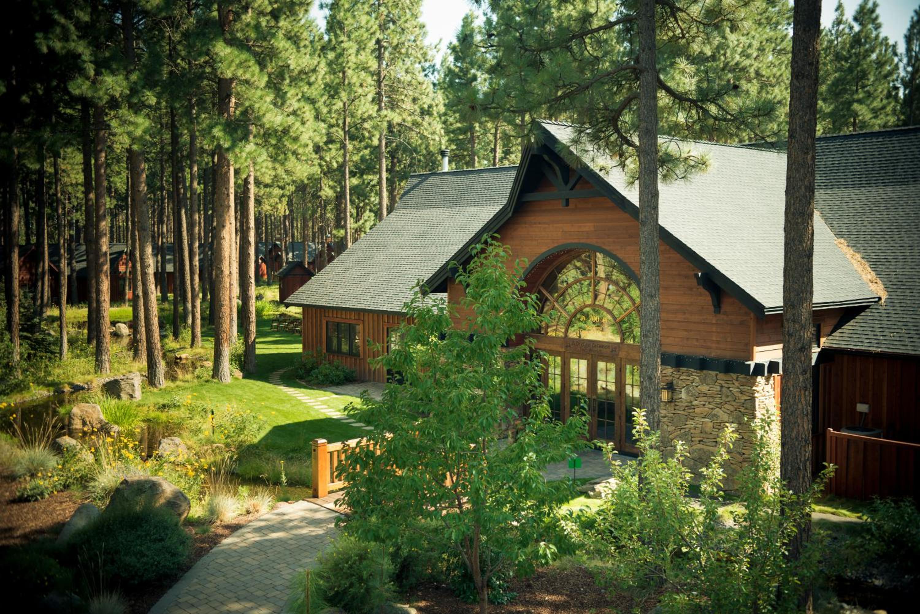 A stone pathway leads past a lawn and pond to a timber lodge featuring an arched glass entryway among pine trees.