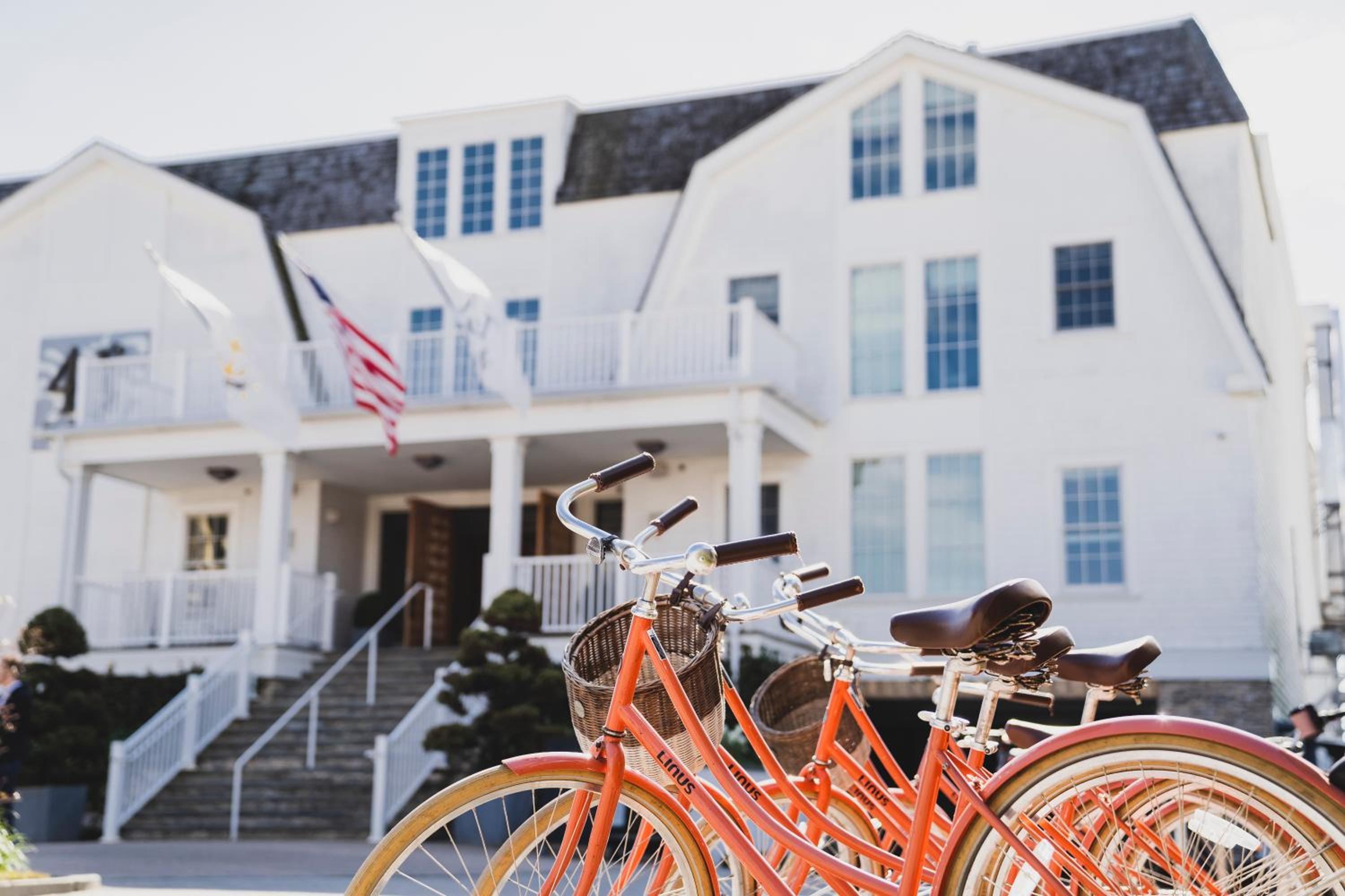 Orange cruiser bicycles with wicker baskets stand before the entrance steps and facade of a white building.
