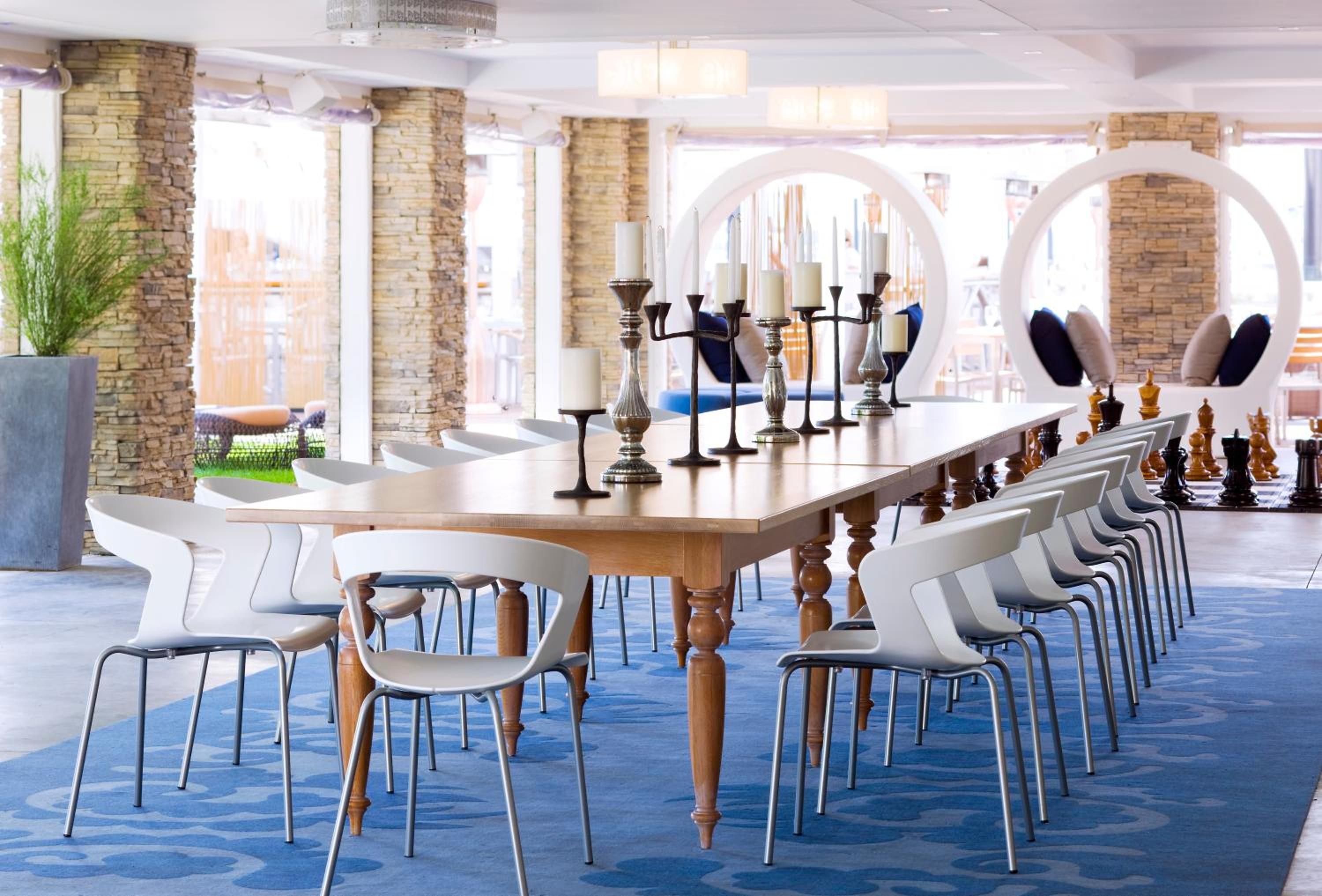 A long wooden table with white chairs and tall candelabras rests on a blue patterned carpet in a bright lounge.